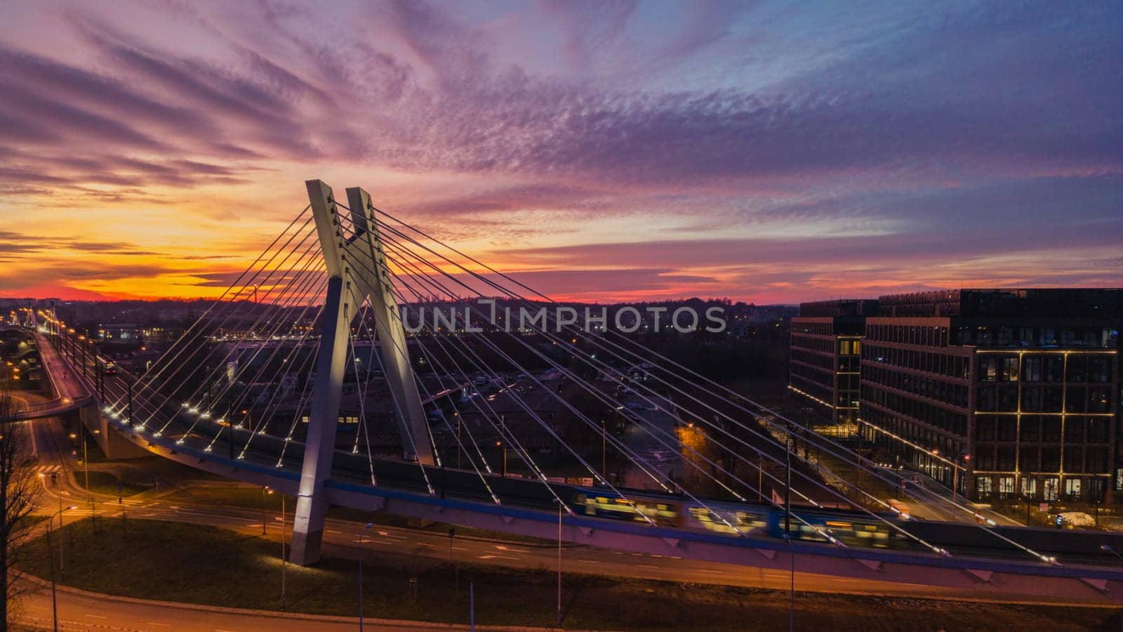 Scenic night aerial cityscape photo with suspension bridge and dramatic dark sunset sky in Krakow, Poland. Perspective above view of high modern bridge structure, street and buildings in lights