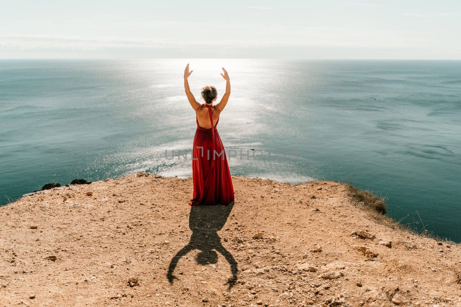 Woman red dress sea. posing on a rocky outcrop high above the sea. Girl on the nature on blue sky background. Fashion photo. by Matiunina