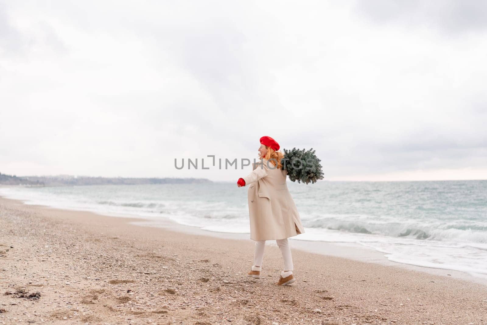 Redhead woman Christmas tree sea. Christmas portrait of a happy redhead woman walking along the beach and holding a Christmas tree on her shoulder. She is dressed in a light coat and a red beret