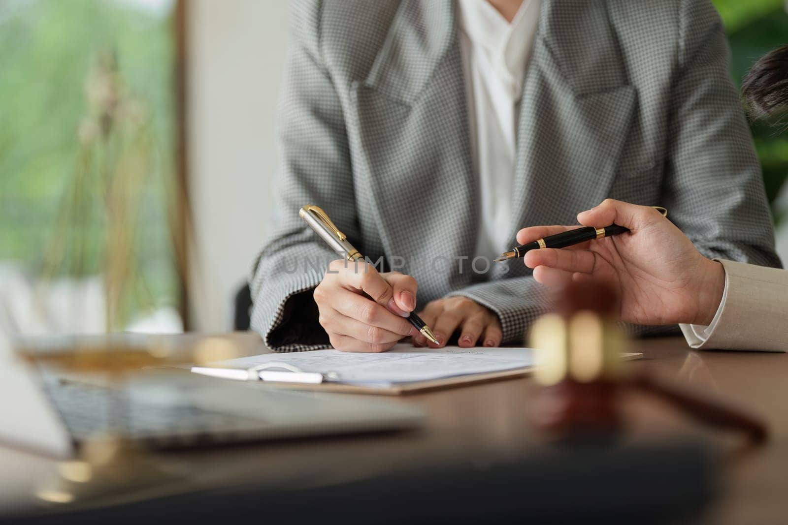 A woman is signing a document at a desk with a laptop and a chair. The woman is wearing a suit and is writing with a pen. The scene suggests a professional setting