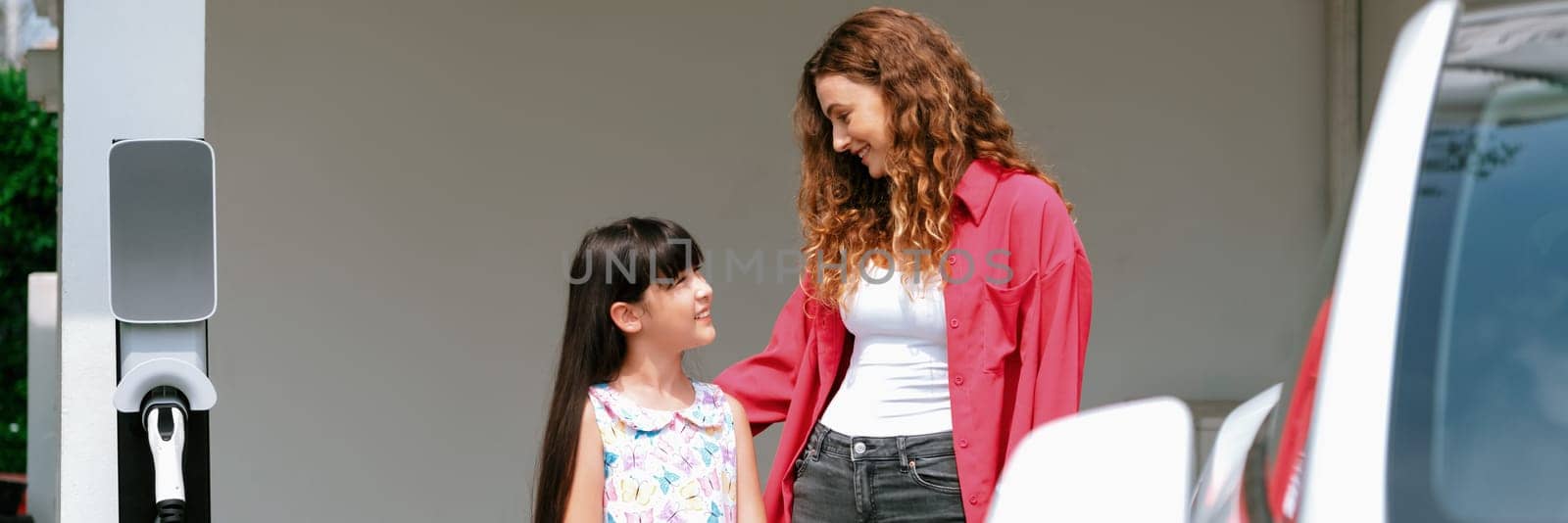 Happy little young girl learn about eco-friendly and energy sustainability as she help her mother recharge electric vehicle from home EV charging station. EV car and modern family. Panorama Synchronos