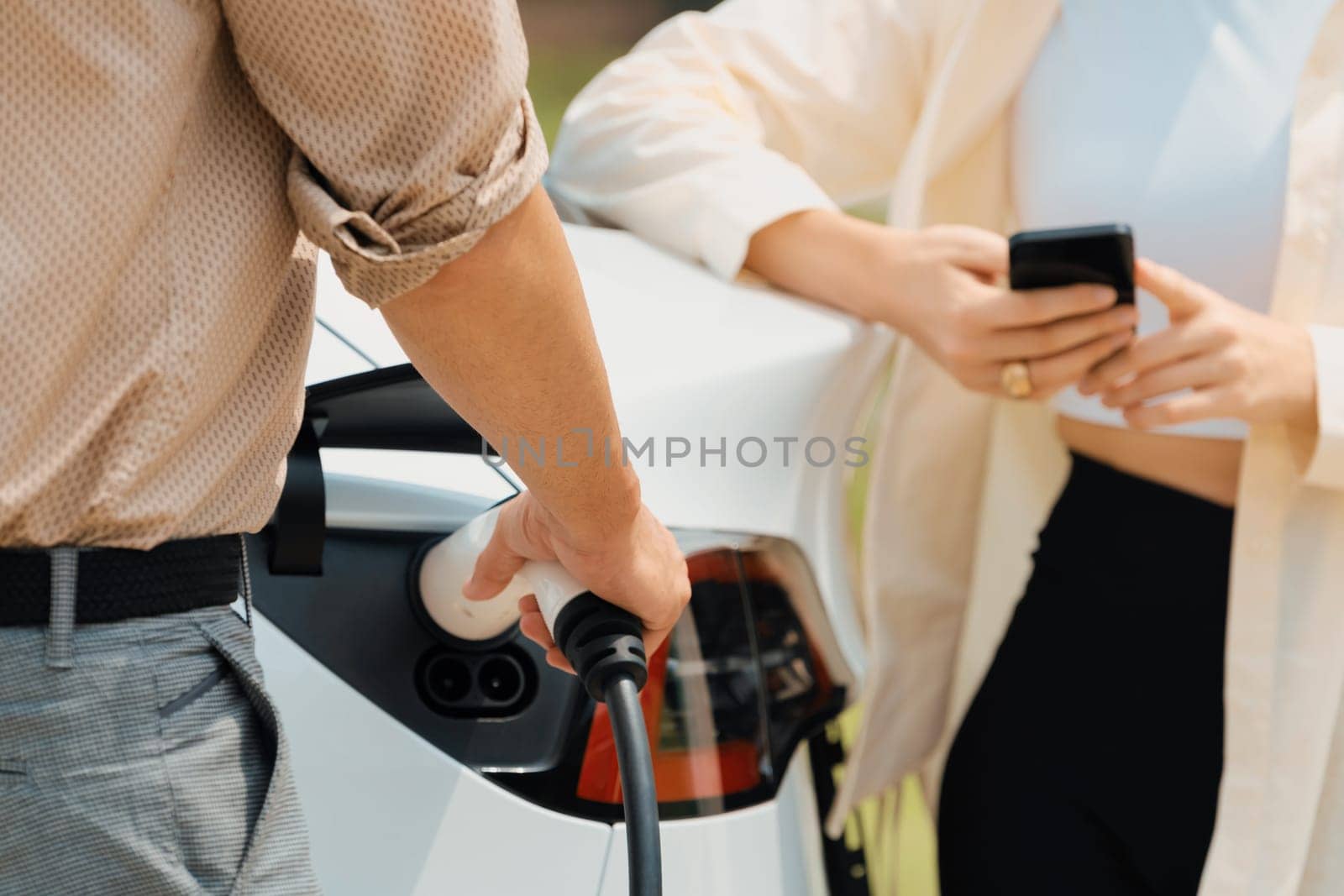 Young couple use smartphone to pay for electricity at public EV car charging station green city park. Modern environmental and sustainable urban lifestyle with EV vehicle. Expedient