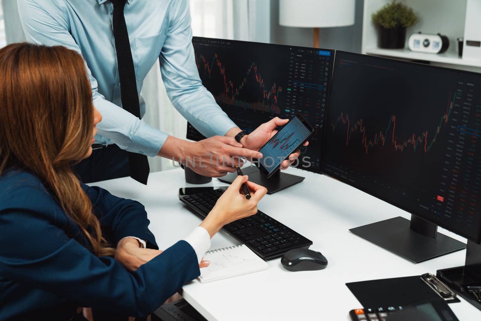 Cropped hand of businessman showing dynamic stock market data on phone to woman writing to memo for analyzing profit value currency rate online website program application at home office. Infobahn.