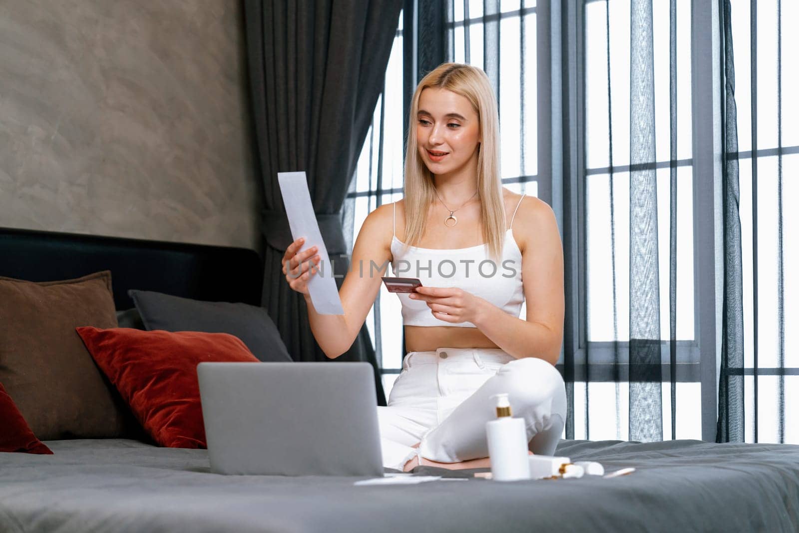 Happy woman sit on the bed smiling as she pays bills with credit card. Blithe by biancoblue