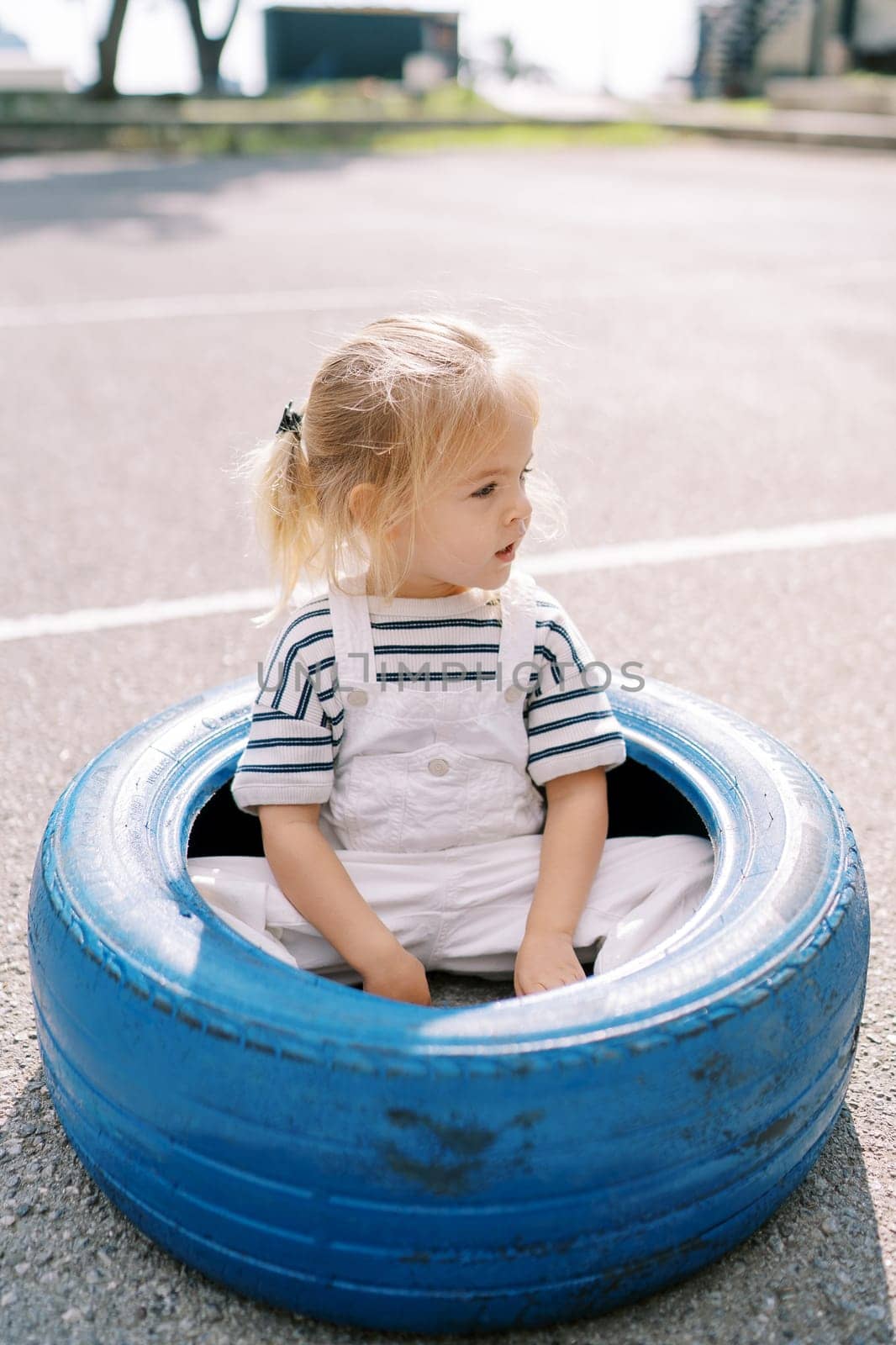 Little girl sits in a big blue tire on the playground and looks away. High quality photo