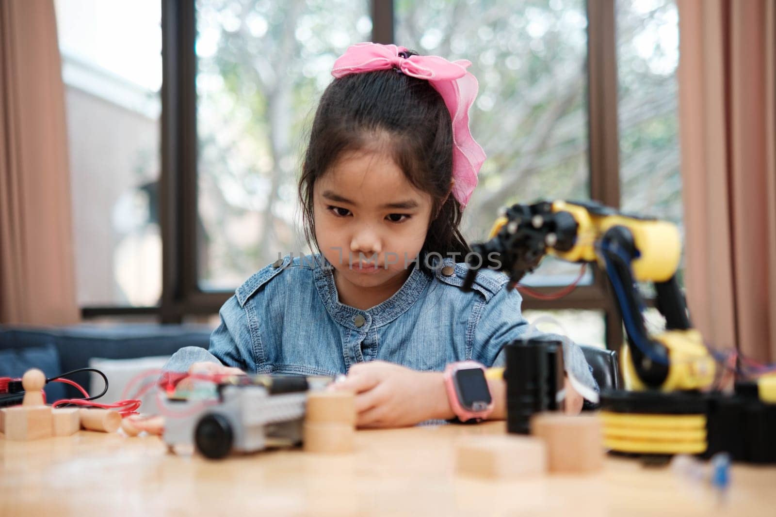 A young Asian girl in a STEM class attentively uses a smartphone app to remotely control a toy car, showcasing tech education.