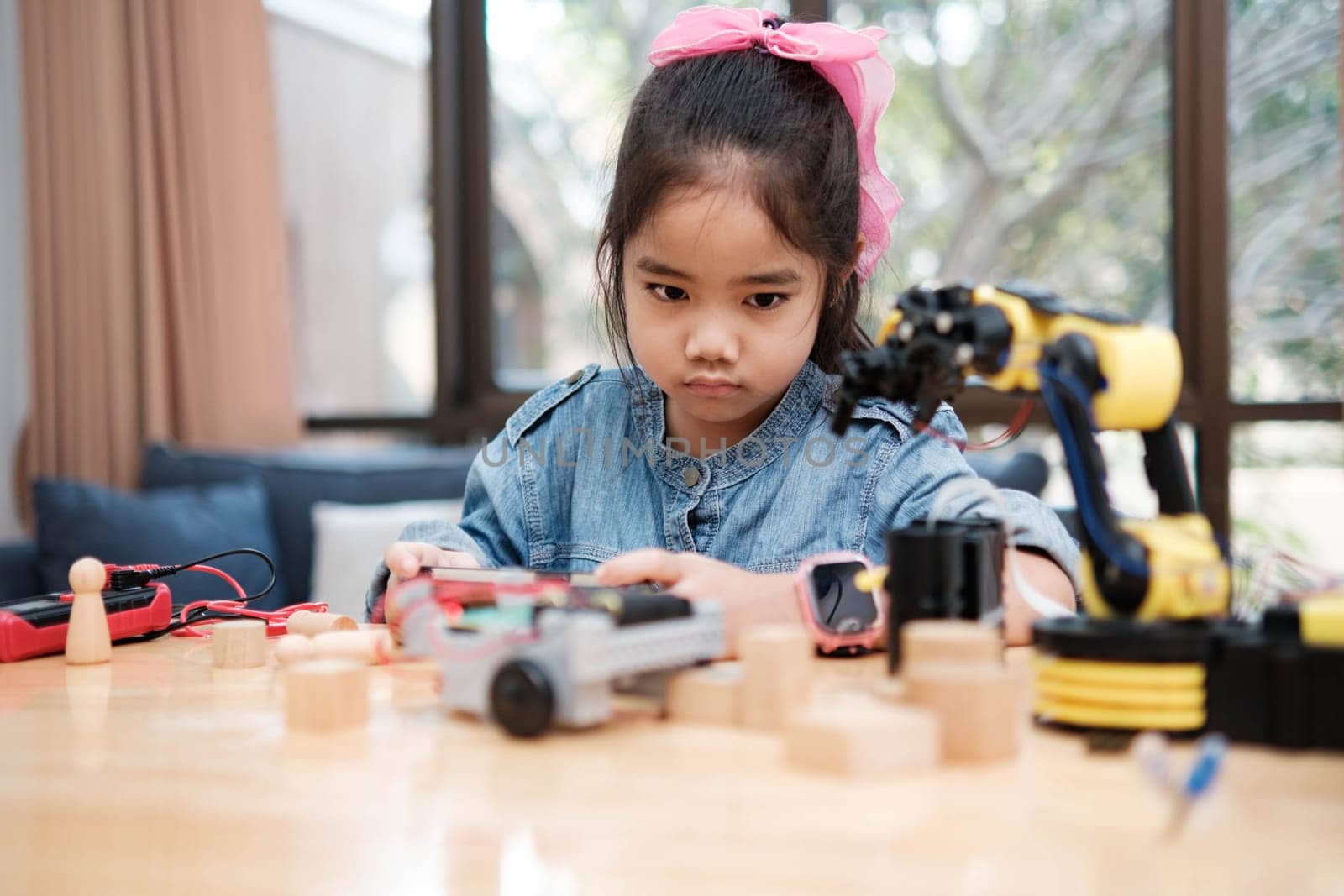 A young Asian girl in a STEM class attentively uses a smartphone app to remotely control a toy car, showcasing tech education.