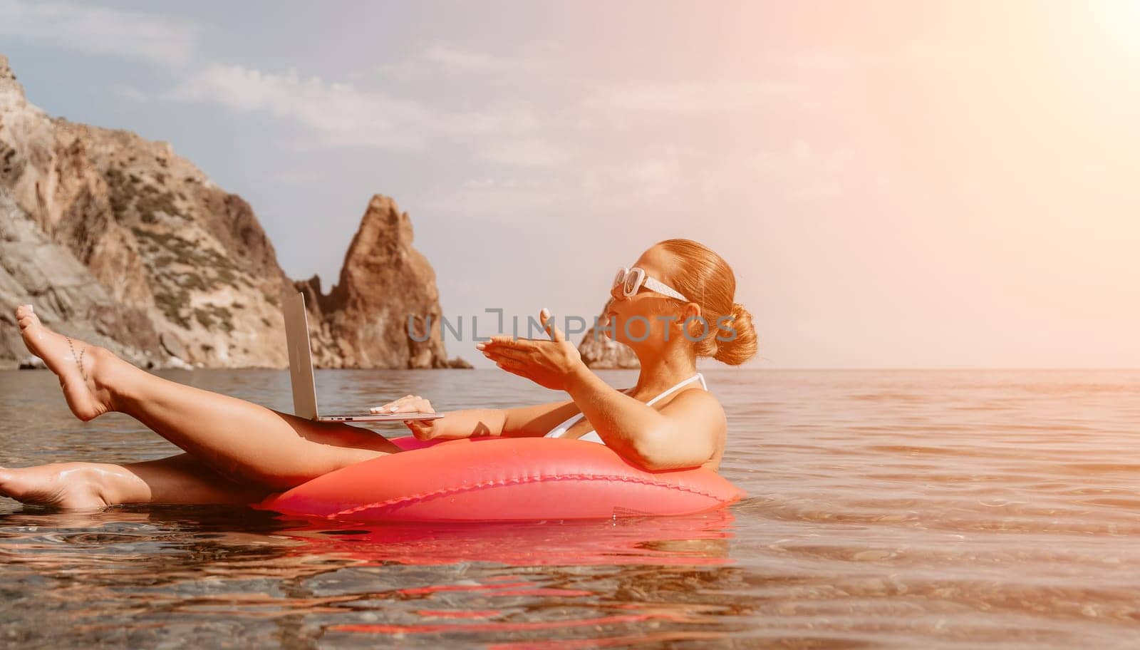 Woman freelancer works on laptop swimming in sea on pink inflatable ring. Pretty lady typing on computer while floating in the sea on inflatable donut at sunset. Freelance, remote work on vacation