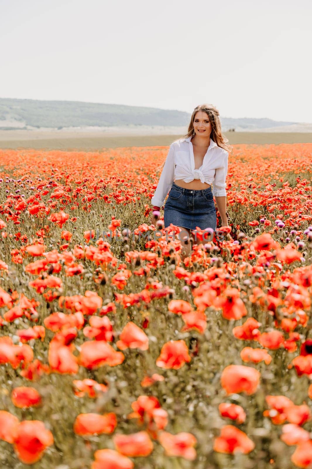 Happy woman in a poppy field in a white shirt and denim skirt with a wreath of poppies on her head posing and enjoying the poppy field