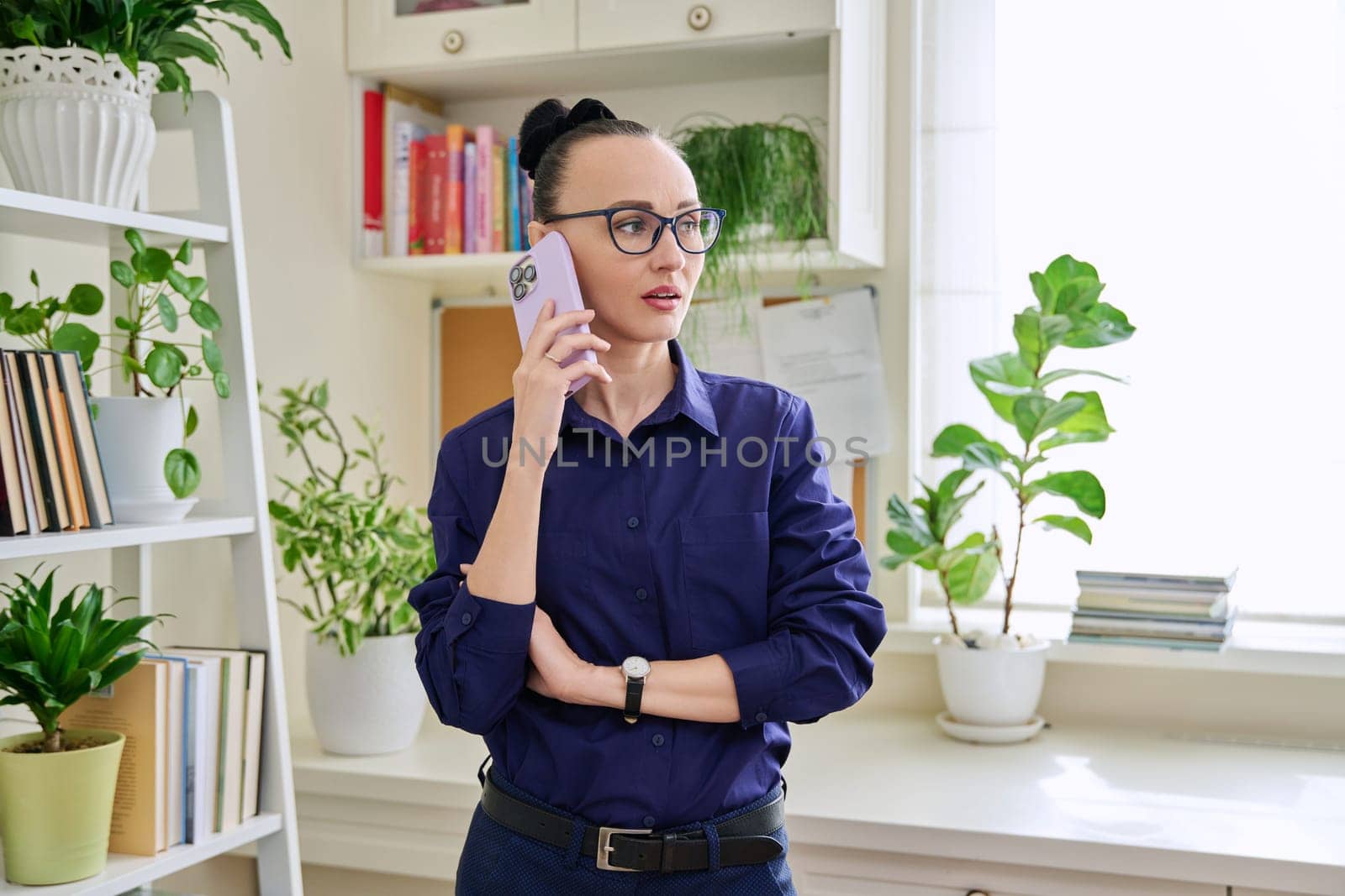 Beautiful emotional woman in her 30s talking on a cell phone, standing in her room at home. Conversation, communication, work, leisure, lifestyle, people concept