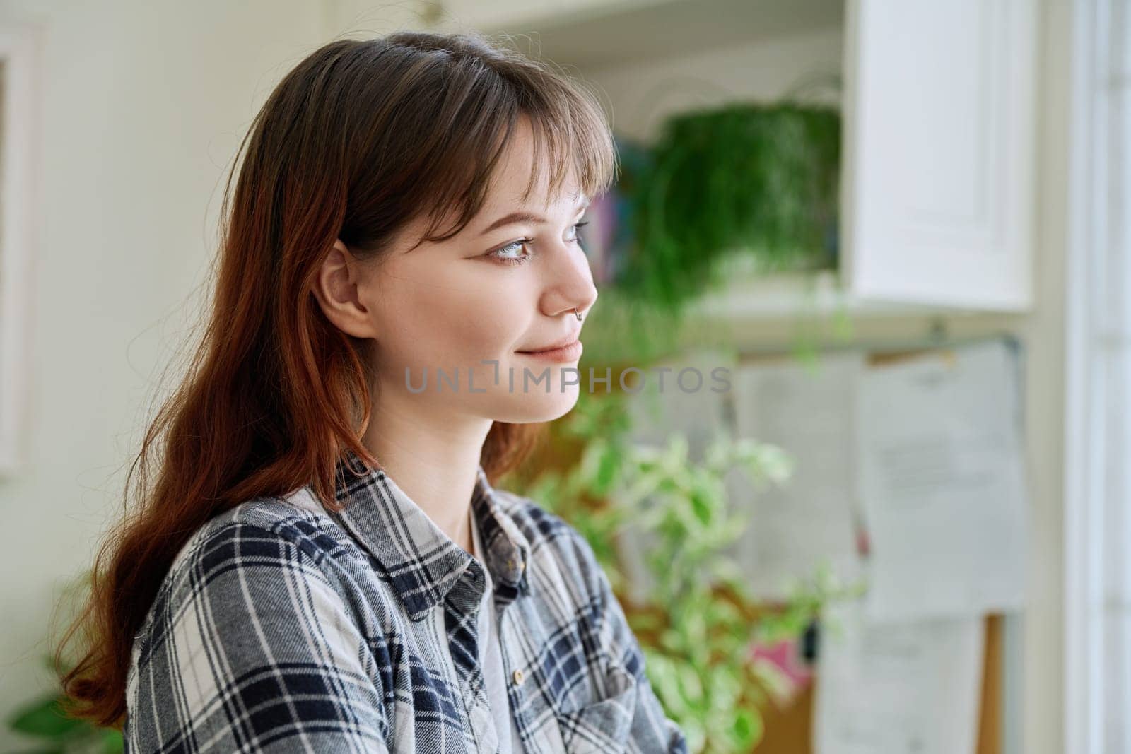 Smiling young woman looking out the window, in home interior, thinking dreaming worrying female, profile view. Lifestyle, feelings, mental health, lifestyle, youth concept