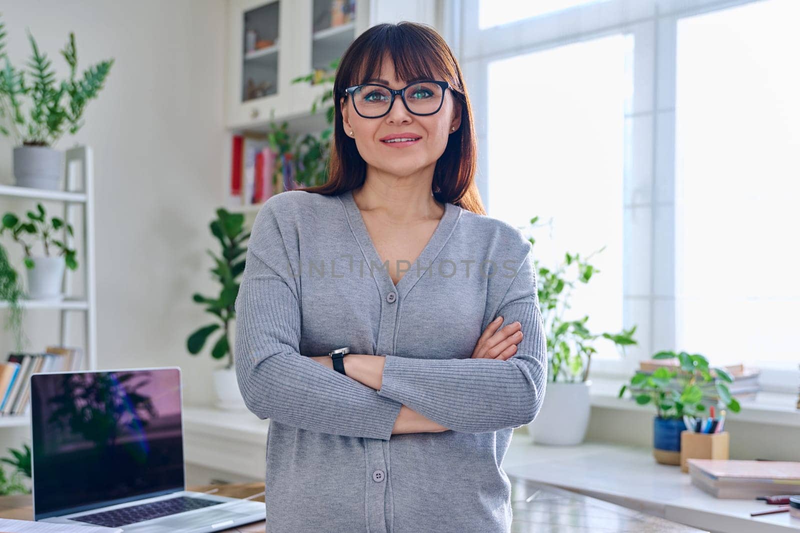 Portrait of confident smiling middle-aged woman in glasses, casual clothes with crossed arms, looking at camera in home interior. 40s age, lifestyle, health, mature lady concept