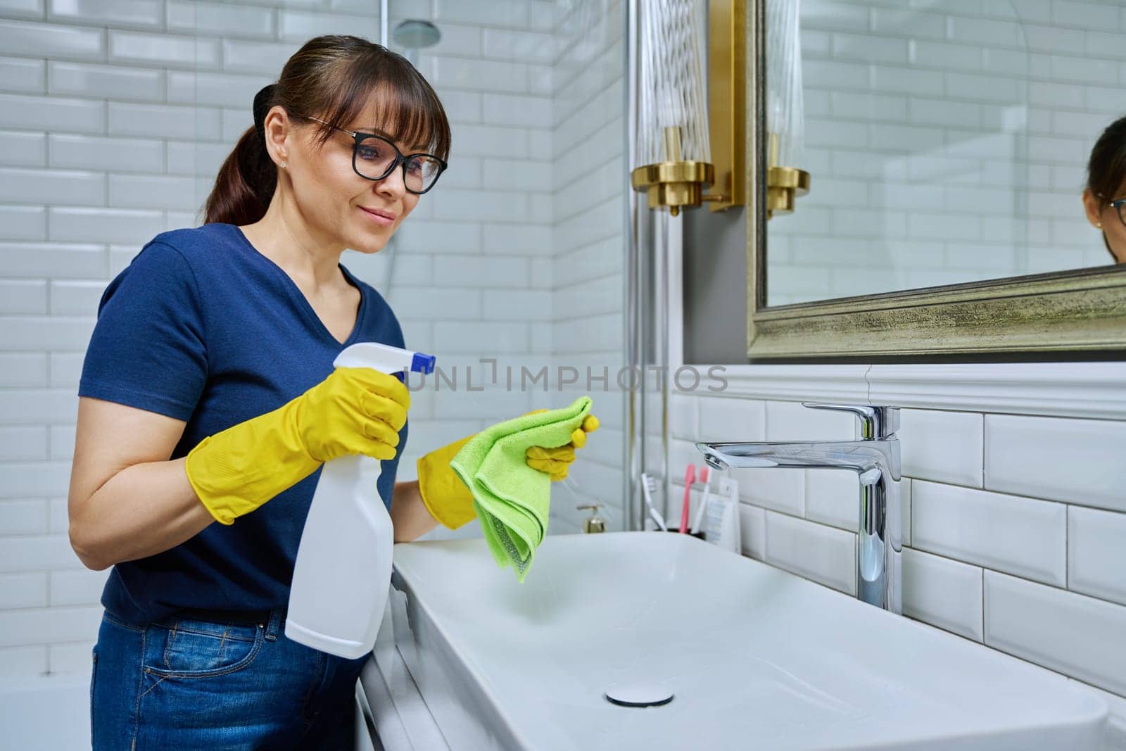 Smiling woman with detergent spray, washcloth cleaning in bathroom, washing sink washbasin. Routine house cleaning, home hygiene, housecleaning service, housekeeping, housework concept