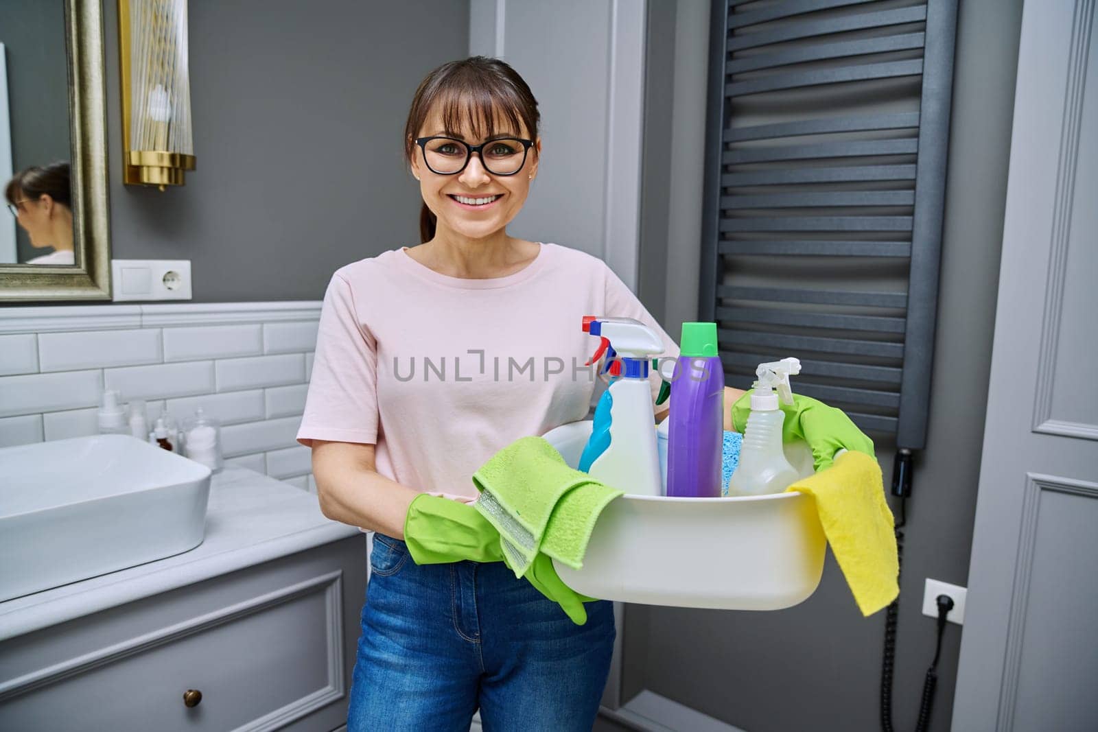 Middle-aged woman in gloves with basin of detergents in bathroom. Female preparing for routine house cleaning, housecleaning service worker posing at workplace. Housekeeping housework cleaning concept