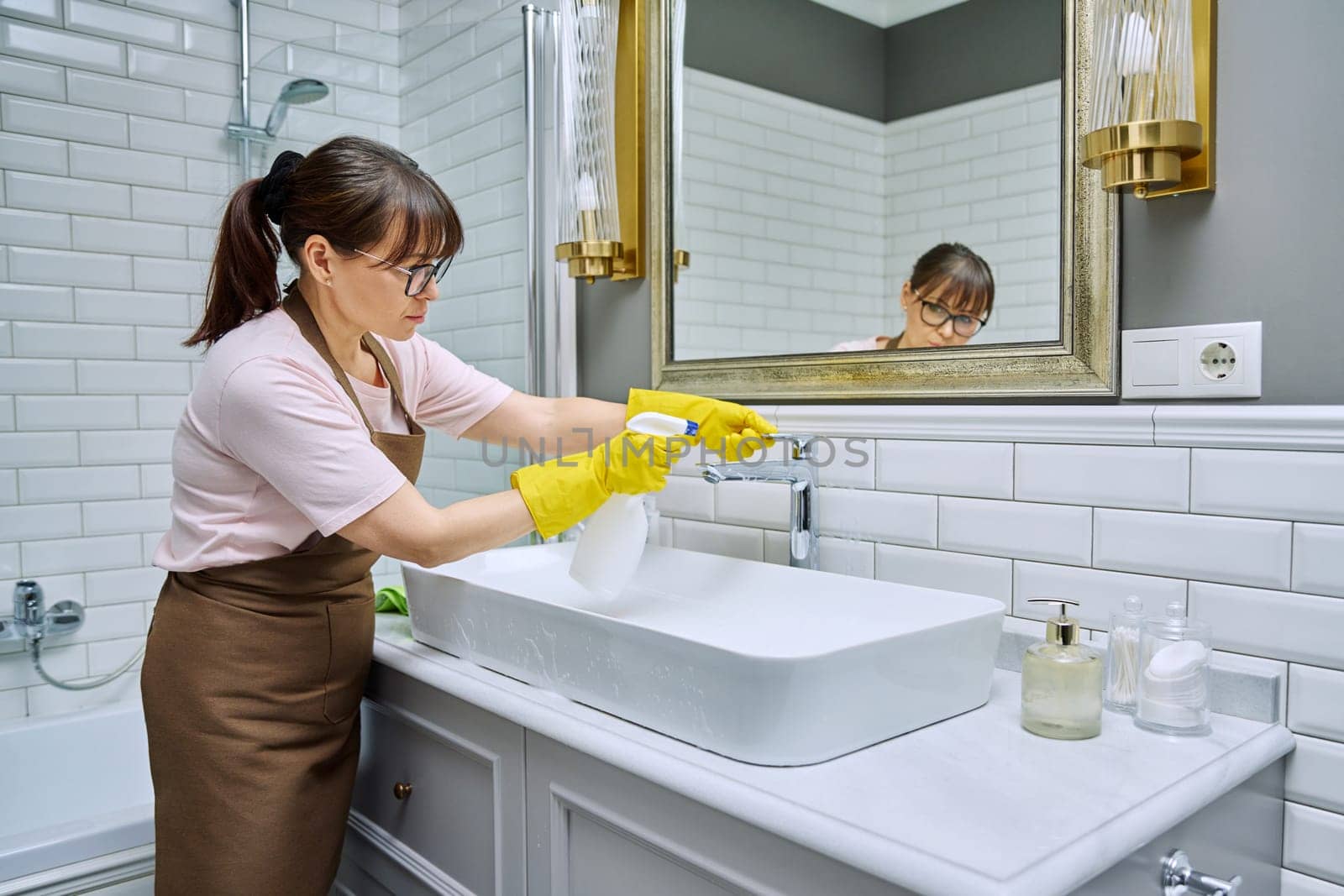 Woman in apron with detergent washcloth cleaning in bathroom, washing sink washbasin. Housewife cleaning house, cleaner service worker at workplace. Home hygiene housecleaning housekeeping housework