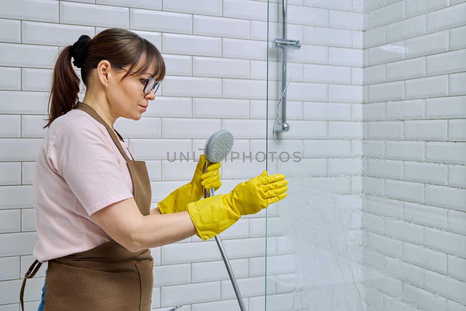 Middle-aged woman in apron, gloves cleaning bathroom, cleans glass in shower. Female housewife cleaning house, service worker at workplace. Home hygiene, housecleaning service, housekeeping, housework
