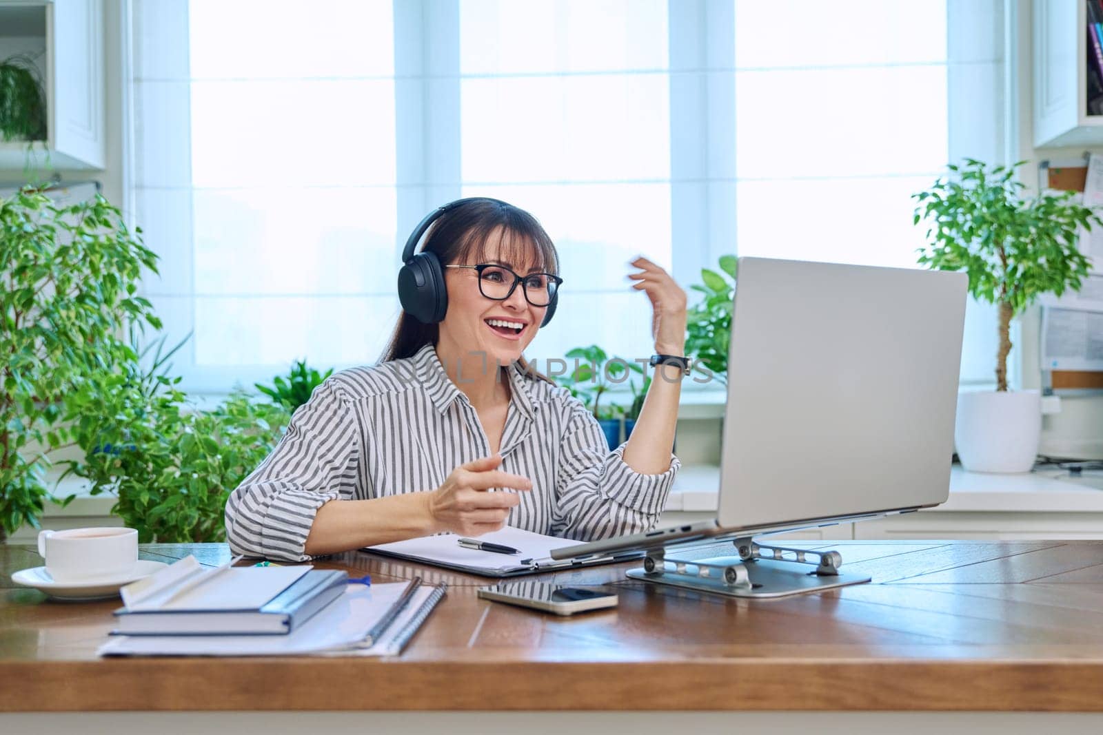 Mature woman in headphones having video conference using laptop computer in home office. Remote virtual meeting, online training, consulting, testing. Work, education, technology, people concept