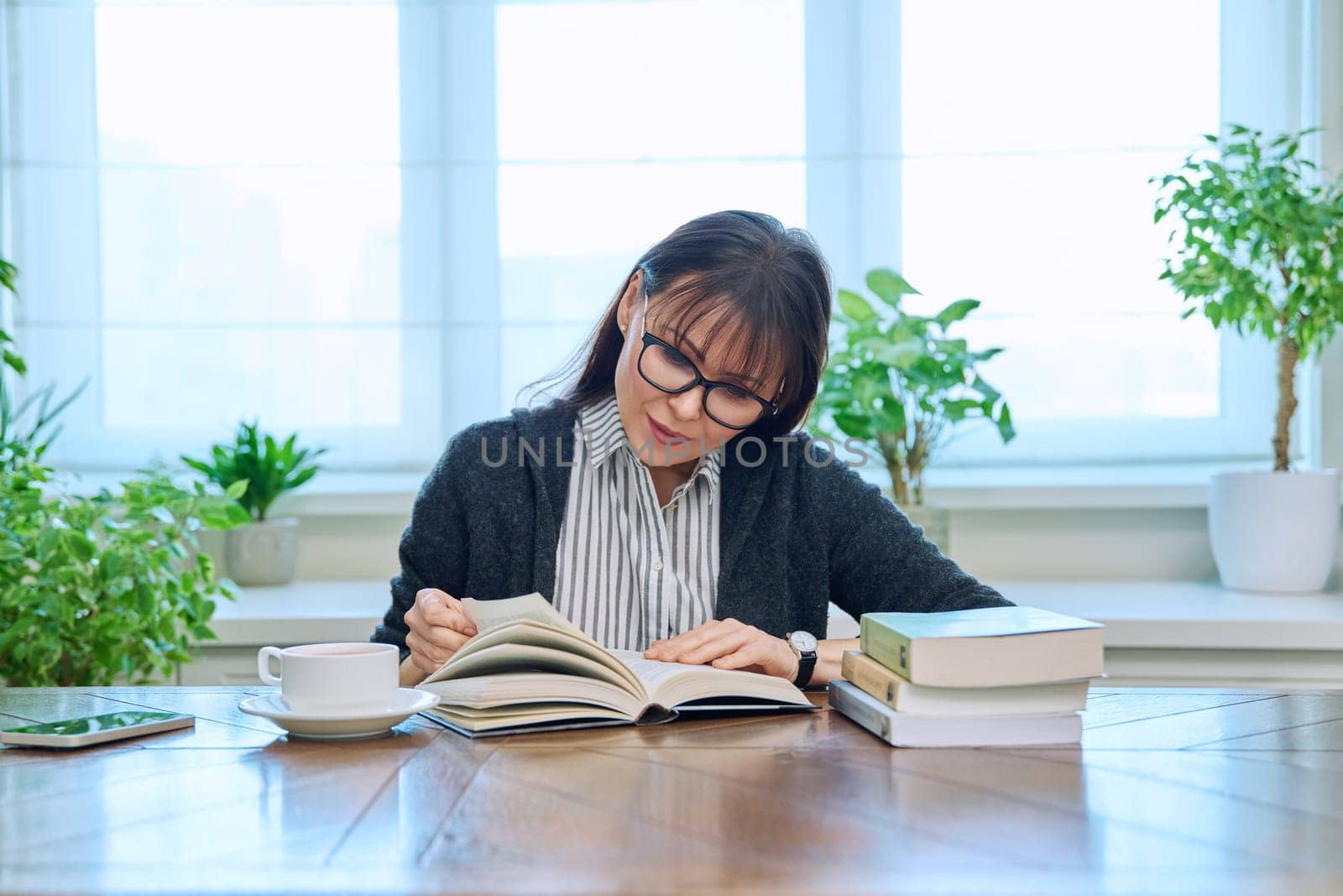 Middle-aged woman reading books, sitting at the table at home. Literary hobby, leisure, relaxation, people concept