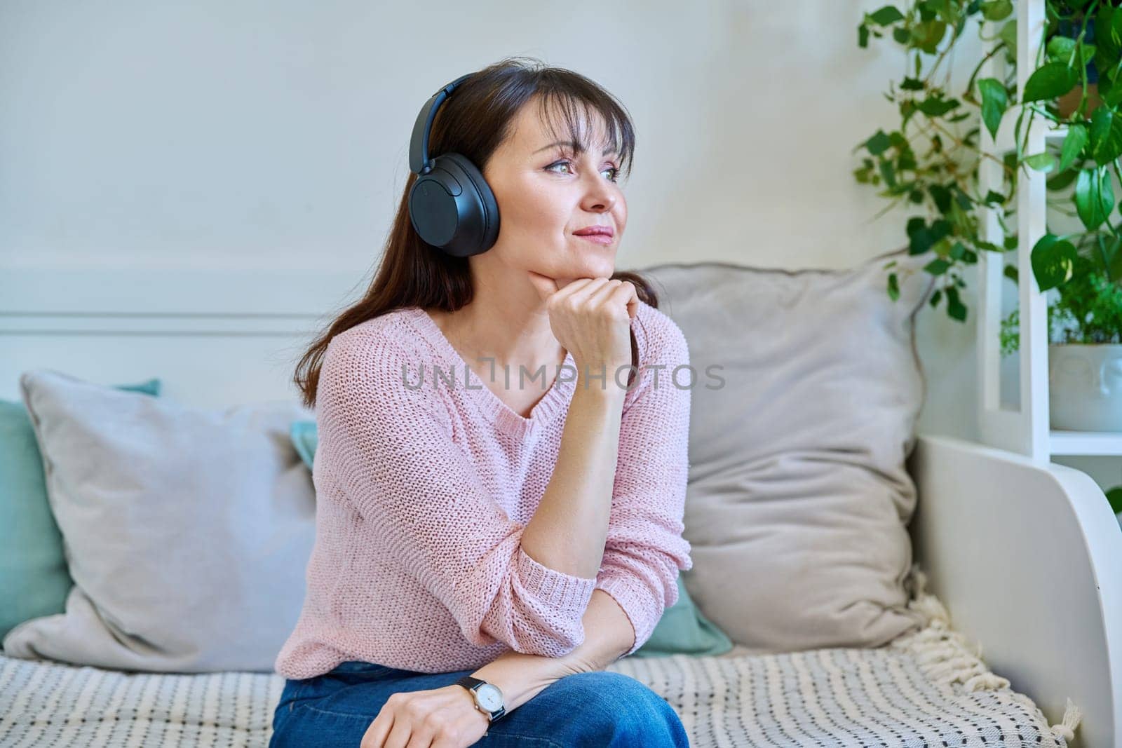 Middle-aged woman in headphones listening to audio, music, audiobook, communication, relaxed vacationer sitting on couch at home