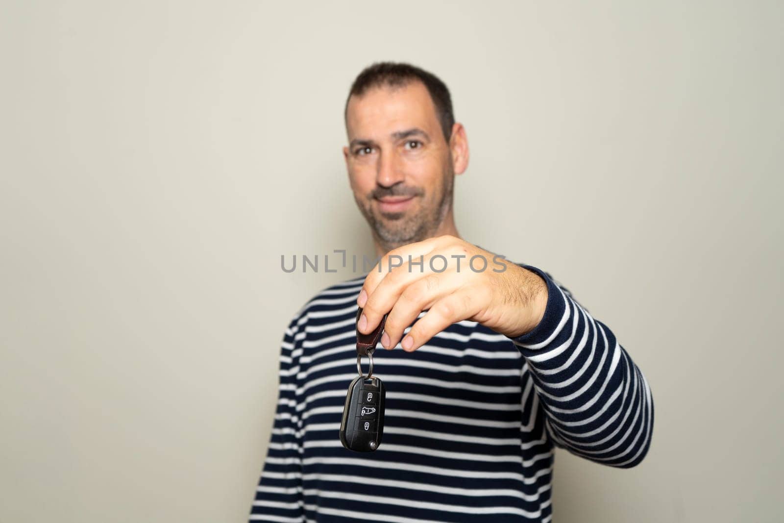 Bearded Hispanic man in his 40s wearing striped sweater, hold car key, keyless system, look at camera isolated on plain beige background studio portrait. People lifestyle concept. Selective focus
