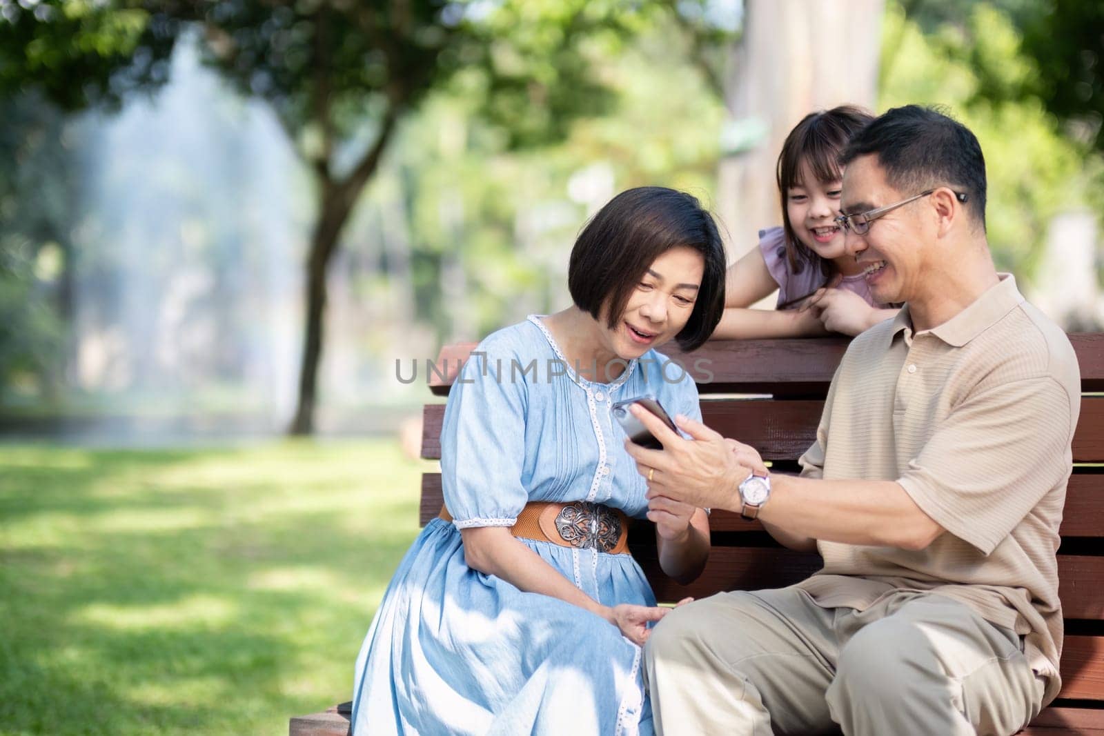 Family, generation and technology concept happy smiling grandmother, grandfather and little granddaughter with telephone sitting at park.