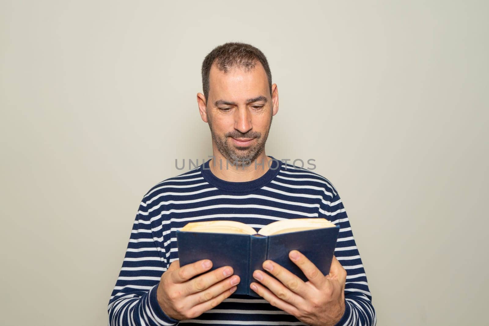 Hispanic man with a beard in his 40s wearing a striped sweater reading a paper book, he is focused and excited about the magnificent content of the story. Isolated on beige background. by Barriolo82