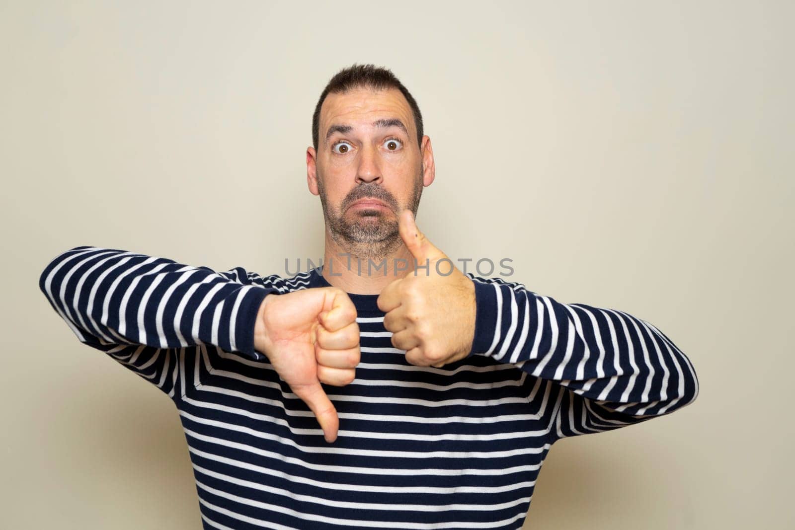 Bearded Hispanic man in his 40s wearing a striped sweater giving a thumbs up and down in doubt and indecision concept, isolated over beige background.