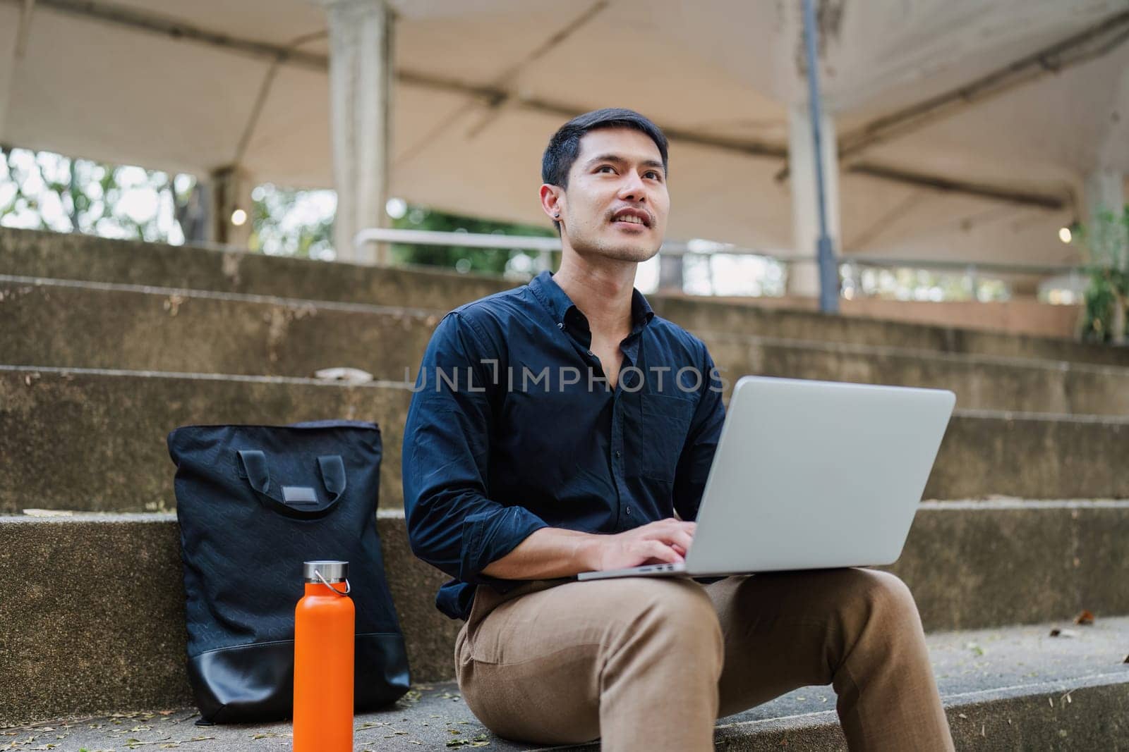 Young businessman working online with laptop while sitting on bench outside of office building.