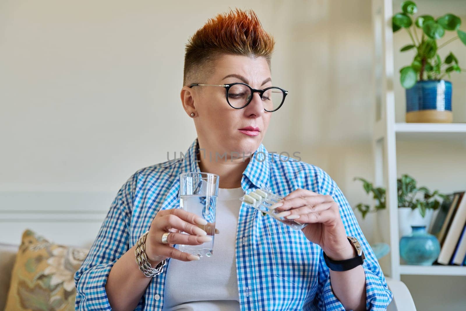 Serious middle aged woman holding blister with capsules glass of water. Female taking painkillers vitamins nutritional supplements antiviral drugs antioxidants antidepressants. Pharmacy health care