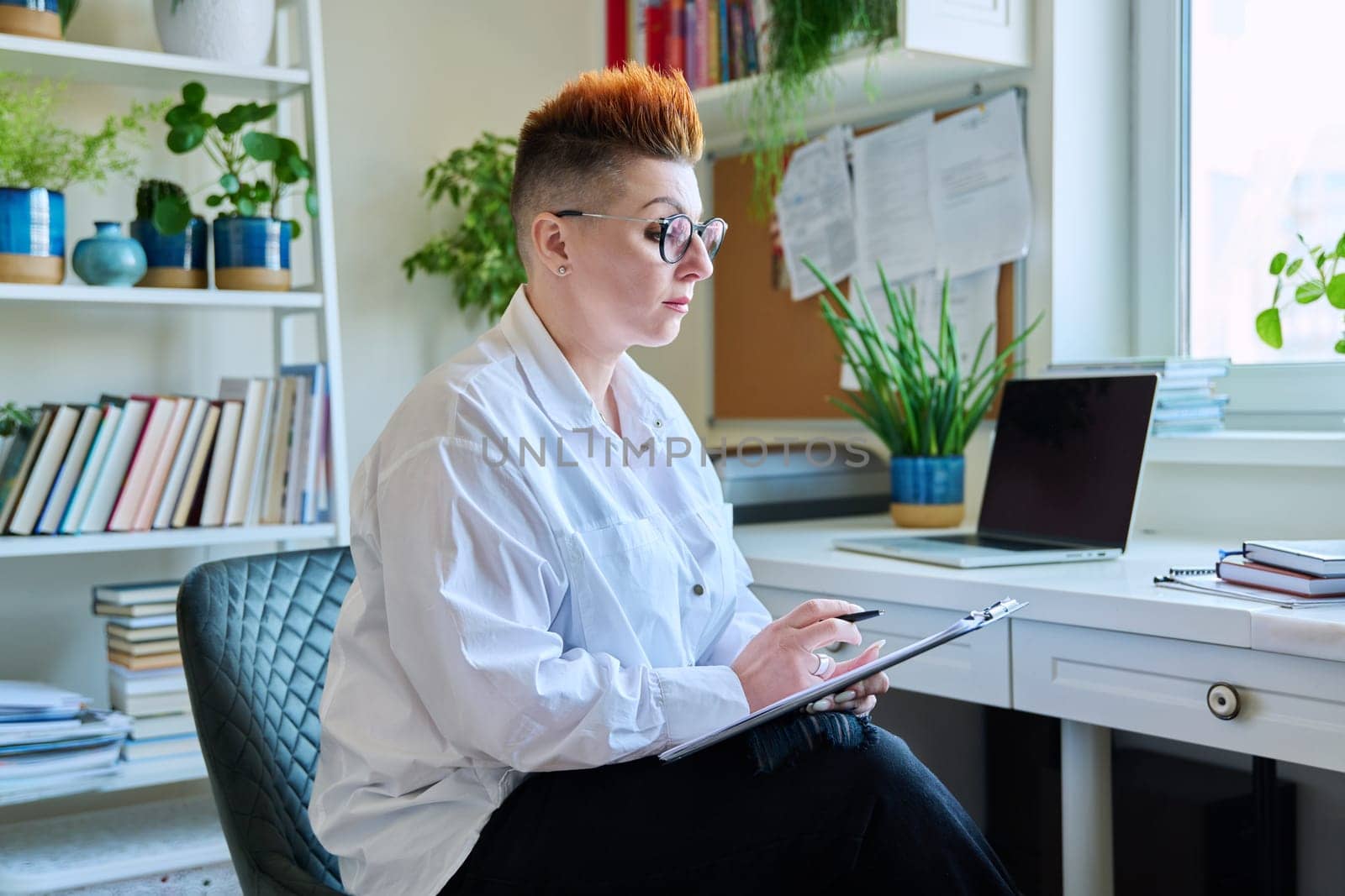 Portrait of serious middle aged female counselor with clipboard at workplace in office by VH-studio