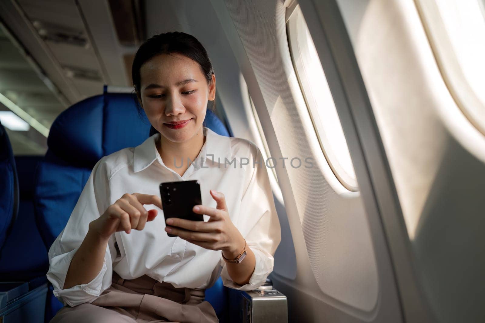 Young Asian woman uses mobile phone to check news information Sitting near the window in business class, airplane class during flight, travel and business concept.