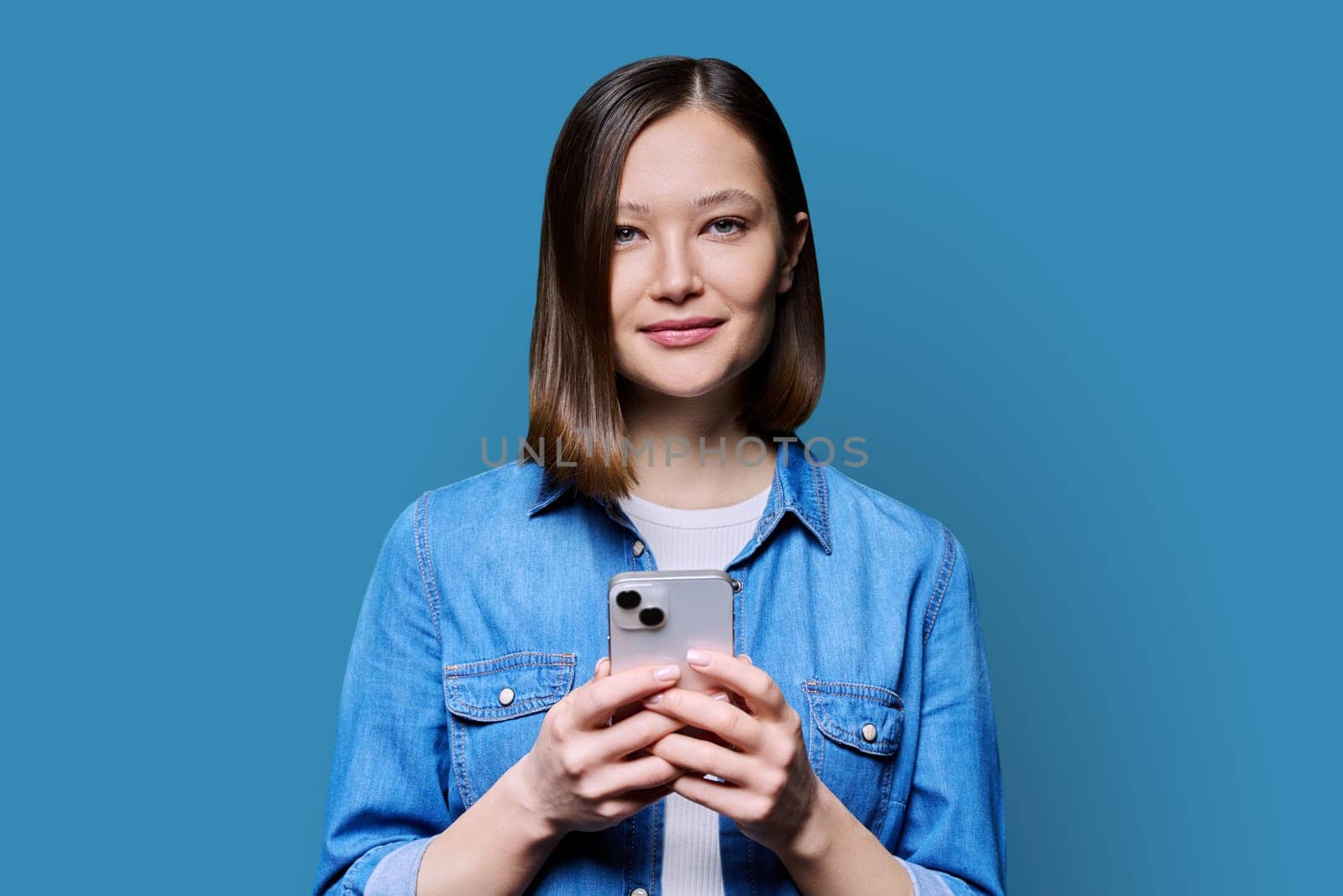 Young happy woman using smartphone in blue background. Smiling 20s female looking at camera texting. Mobile Internet apps applications technologies for work education communication shopping healthcare