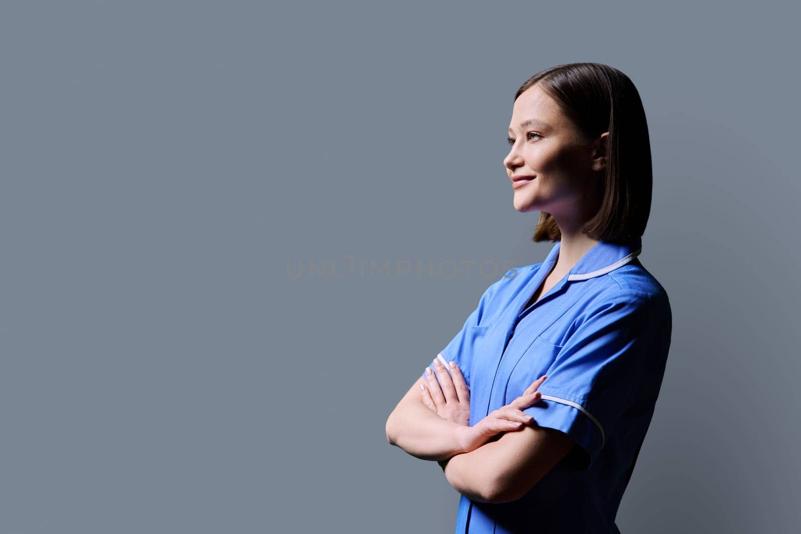 Portrait of young confident smiling female nurse with crossed arms, looking in profile on gray studio background, copy space. Medical services, health, professional assistance, medical care concept