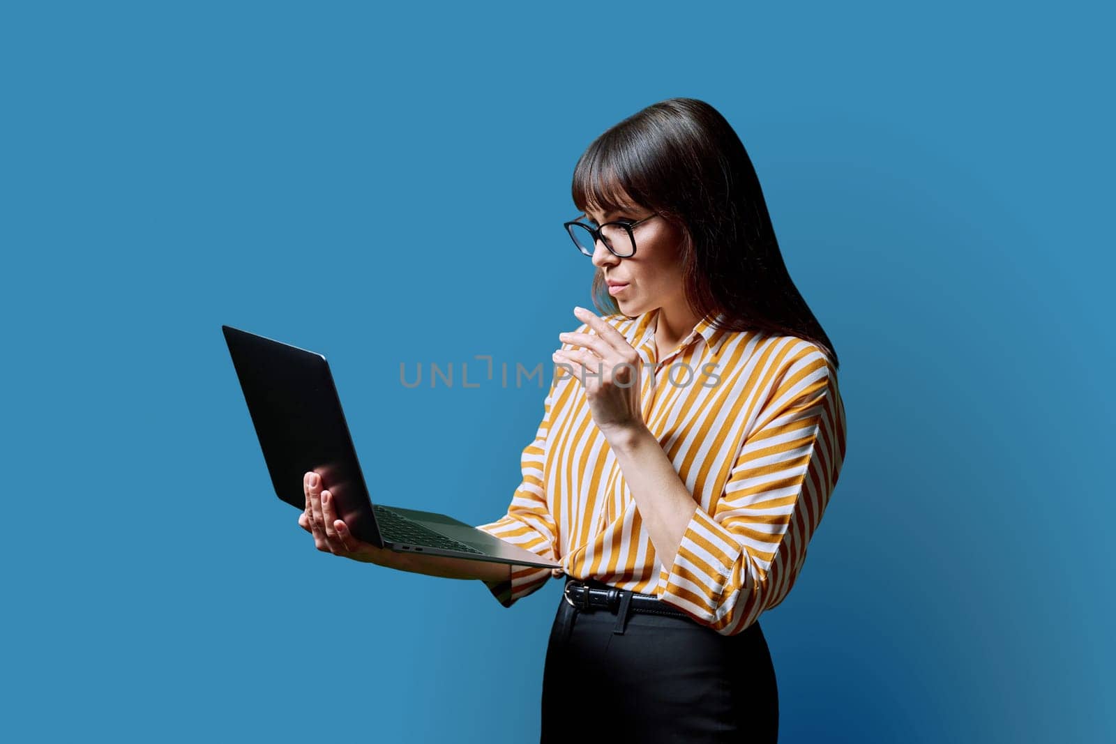 Serious middle aged woman holding laptop on blue studio background. Confident mature female working using computer. Internet online technology, business, work, service, lifestyle