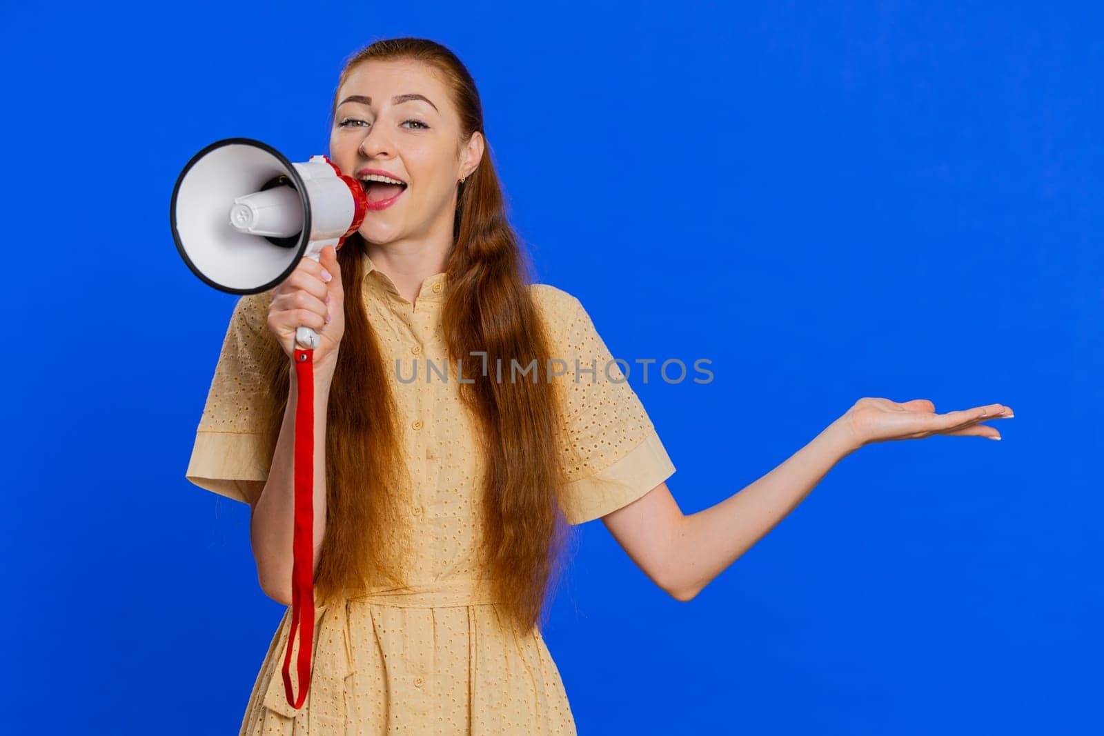 Woman talking with megaphone, proclaiming news, loudly announcing advertisement, warning using loudspeaker to shout speech, pointing empty place. Copy-space. Pretty redhead girl on blue background