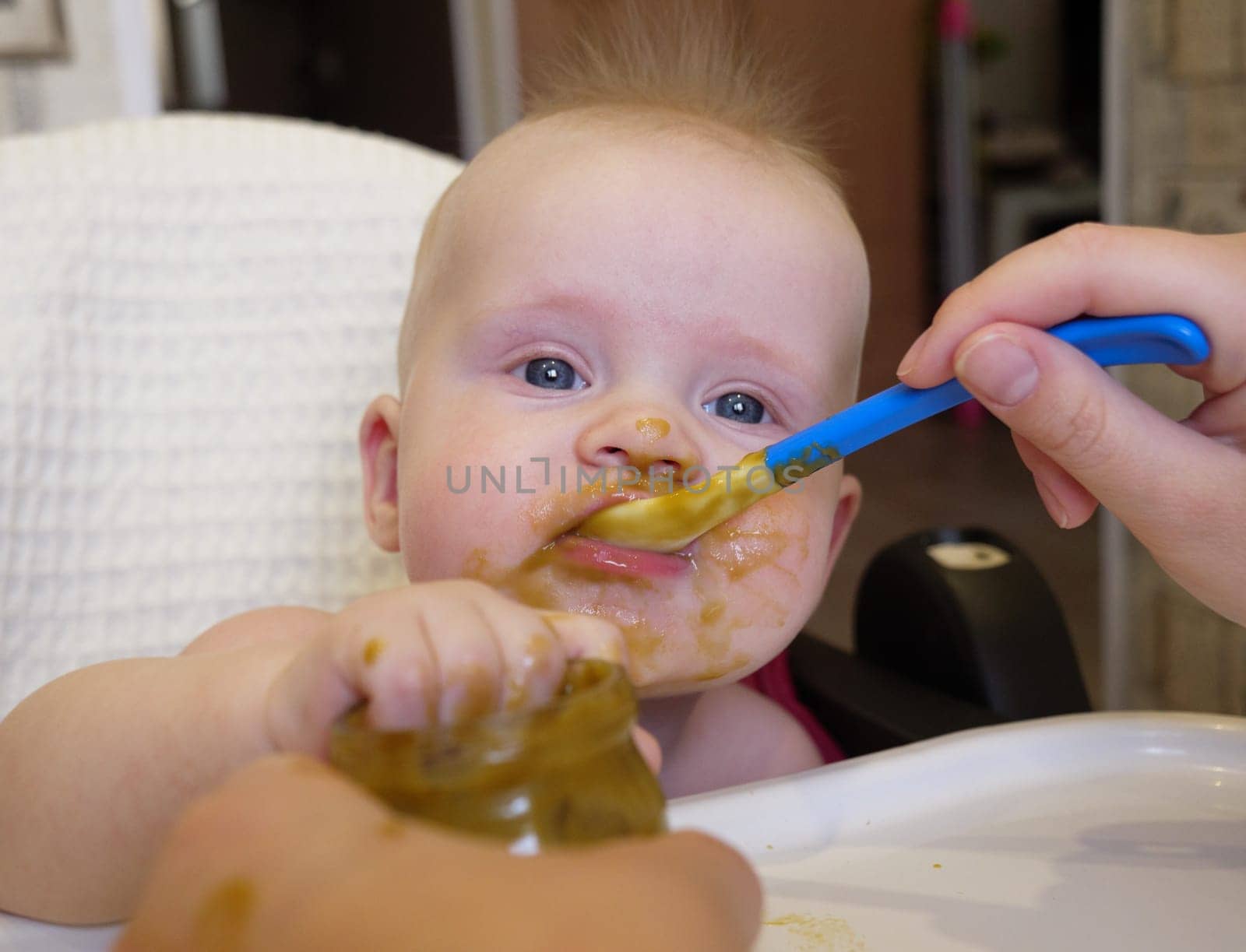 Mom feeding little boy with broccoli puree. Child at the age of six months eats broccoli while sitting on a baby chair.
