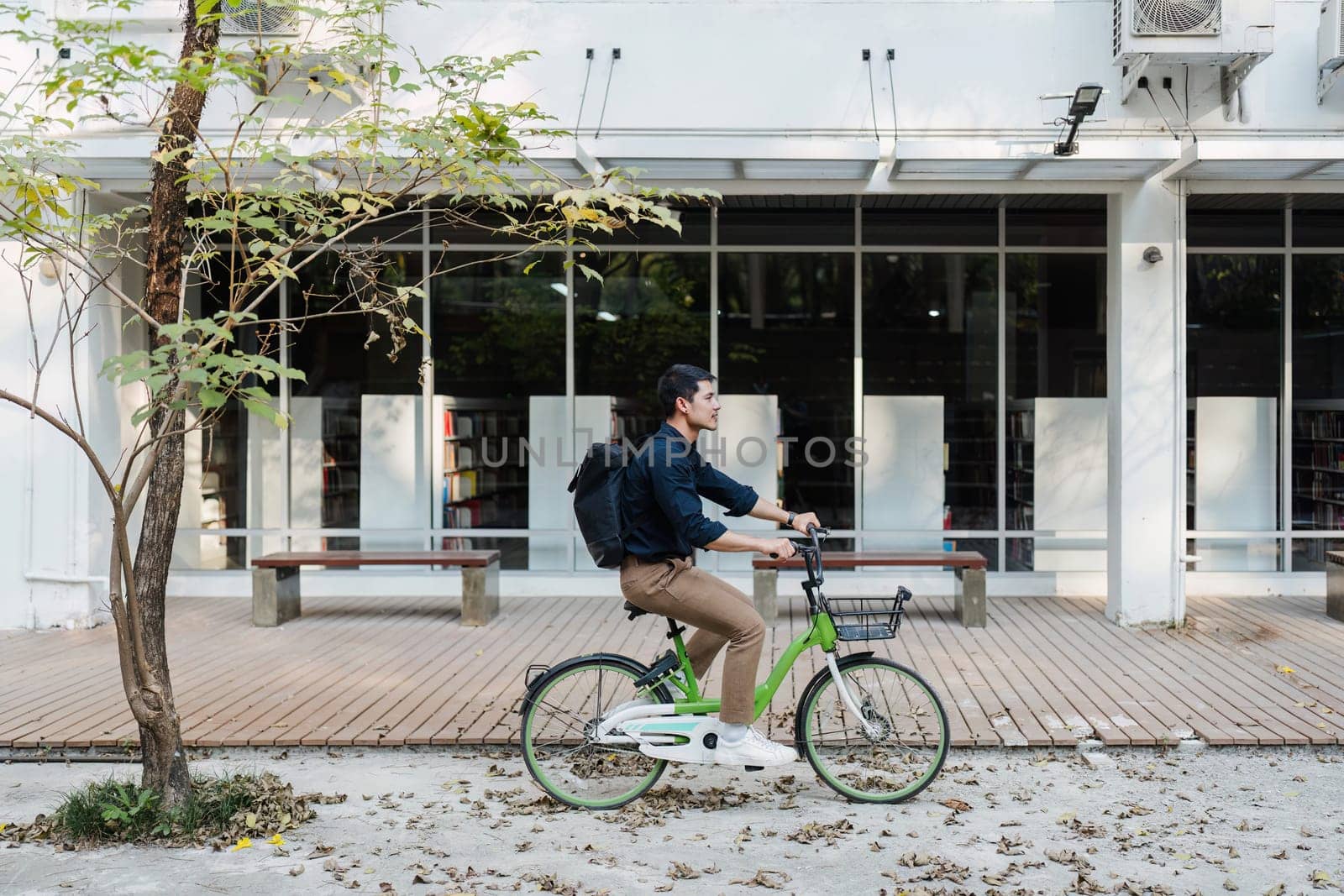 A man rides a bicycle down a sidewalk in front of a building with a lot of books. The man is wearing a backpack and a blue shirt. The scene is peaceful and calm, with the man enjoying his ride