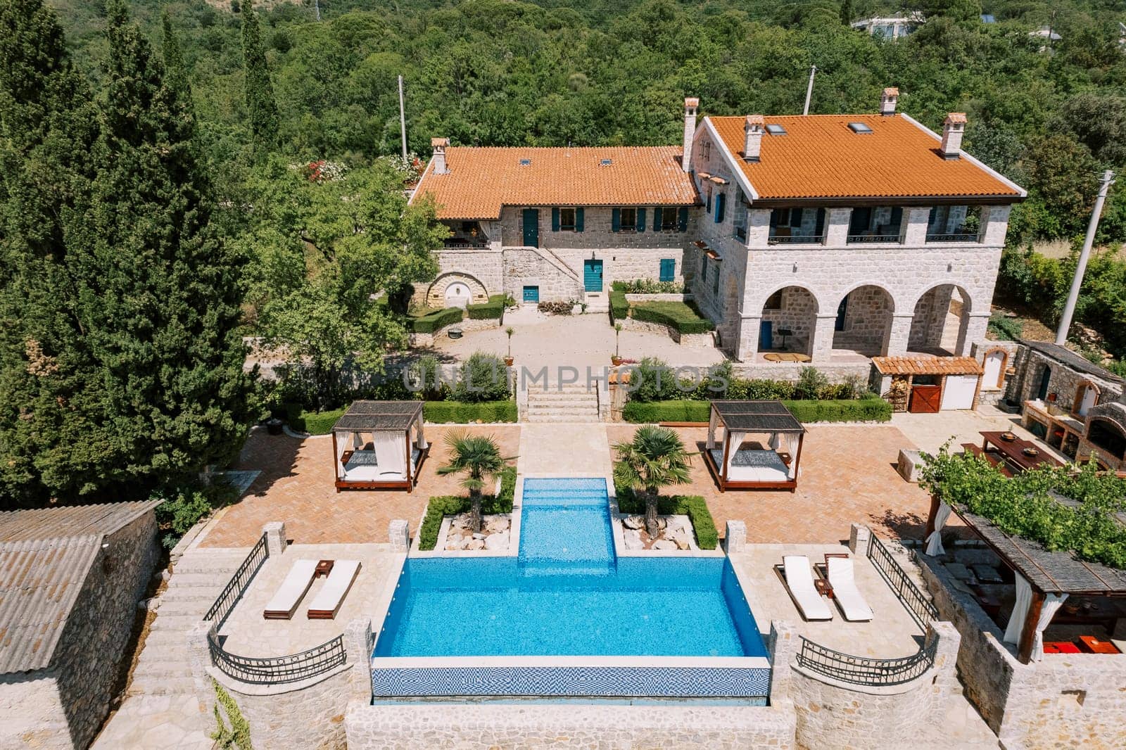 Sun loungers stand near the T-shaped pool in the courtyard of an ancient stone villa in a green garden. Drone. High quality photo