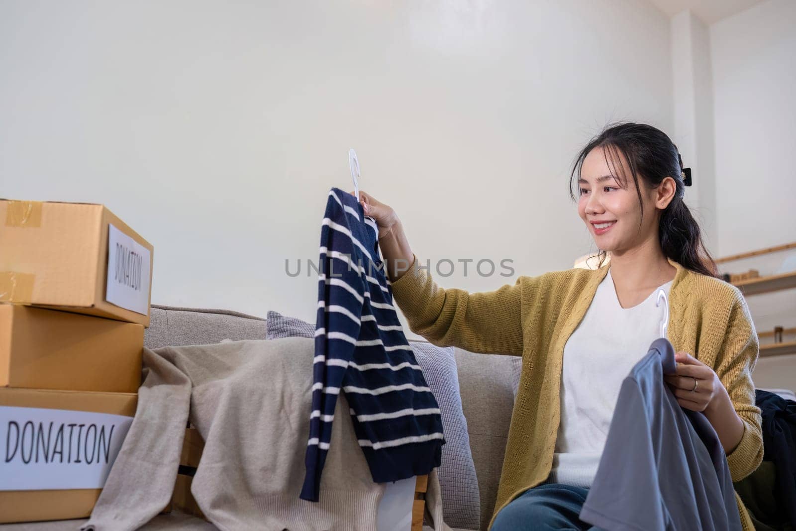 A young Asian woman sits on a sofa sorting unwanted clothes to donate to a charity..