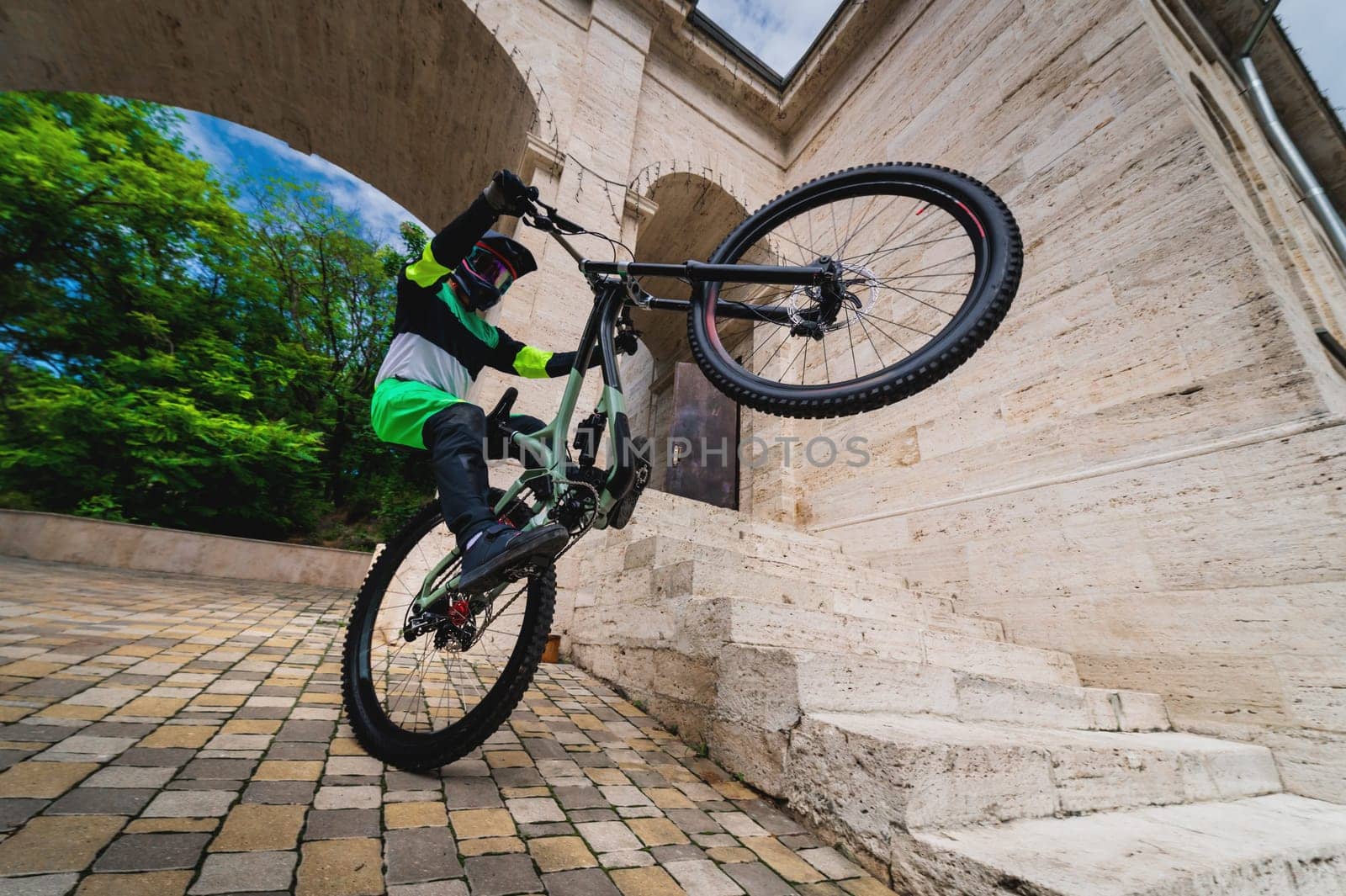 A man standing on one wheel of a mountain bike. Horizontal shot outdoors in the city with architecture in the background, wide angle. Guy performs a sports trick.