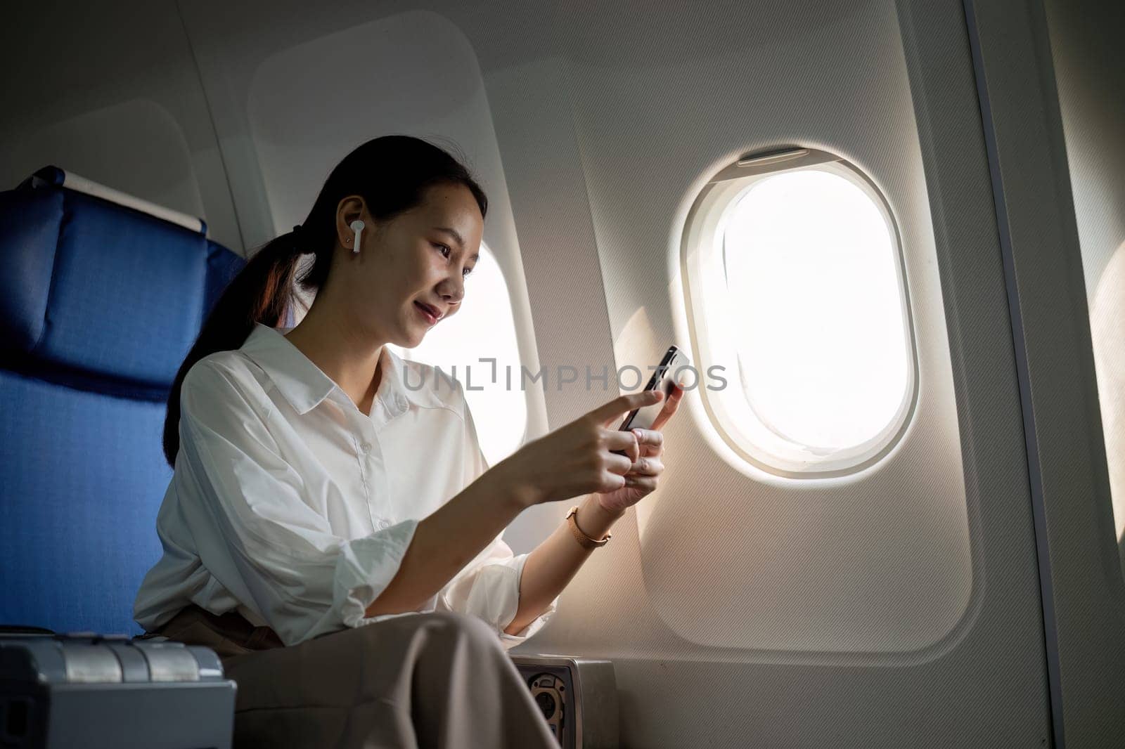 Young Asian woman uses mobile phone to check news information Sitting near the window in business class, airplane class during flight, travel and business concept by wichayada