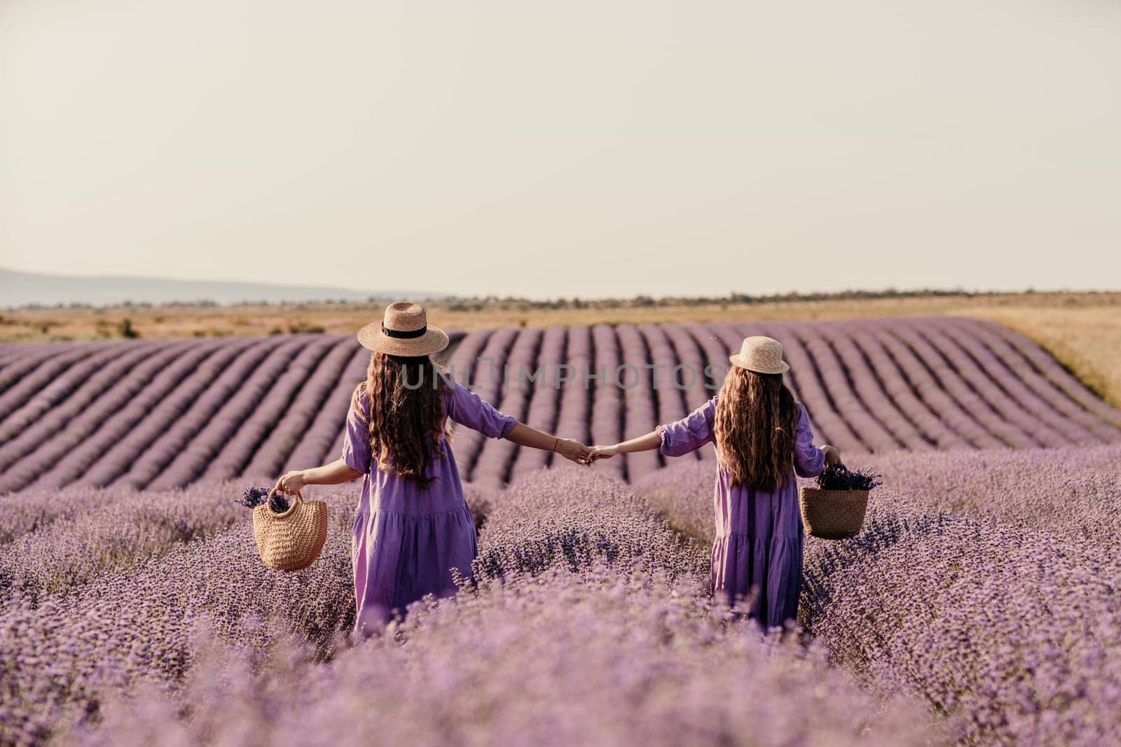 Mom and daughter are running through a lavender field dressed in purple dresses, long hair flowing and wearing hats. The field is full of purple flowers and the sky is clear. by Matiunina