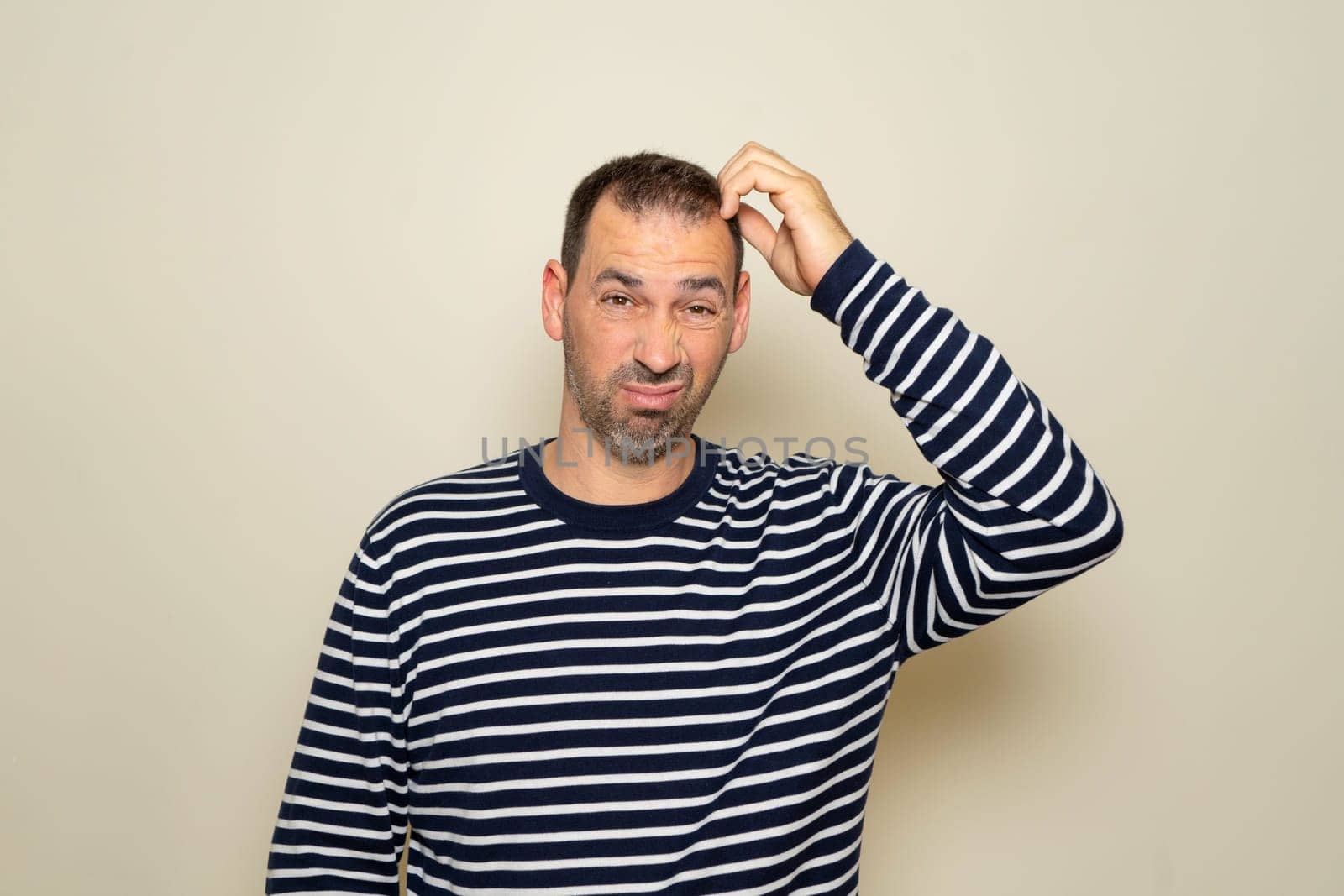 Bearded Hispanic man in his 40s wearing a striped sweater scratching his head while trying to make a decision, doubt concept. Isolated on beige studio background