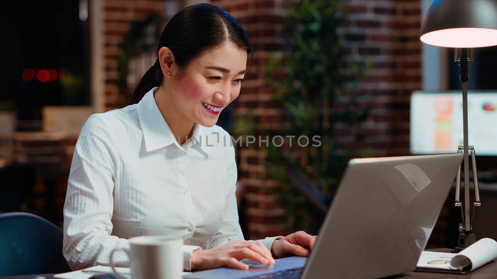 Asian woman dancing in office, feeling excited after learning of upcoming promotion, rewarded for good results. Cheerful employee singing on computer desk chair, celebrating, camera B