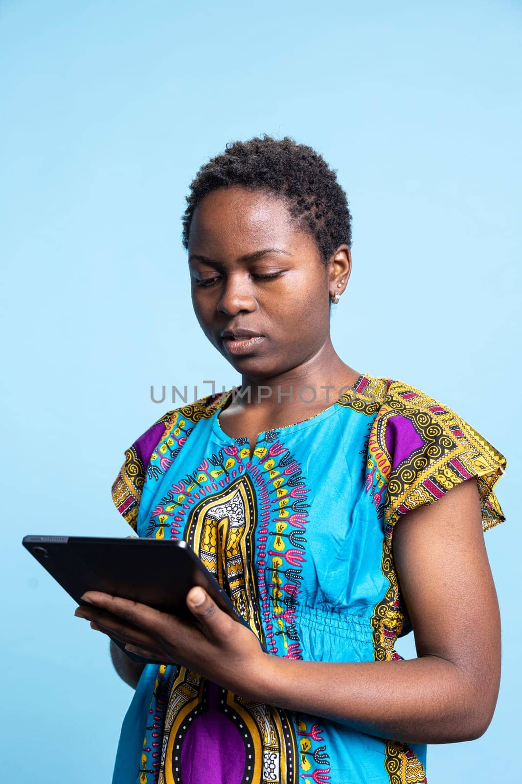 African american woman is using a modern tablet in studio, scrolling through online webpages over blue background. Girl holding device to stay connected with people on social media platforms.