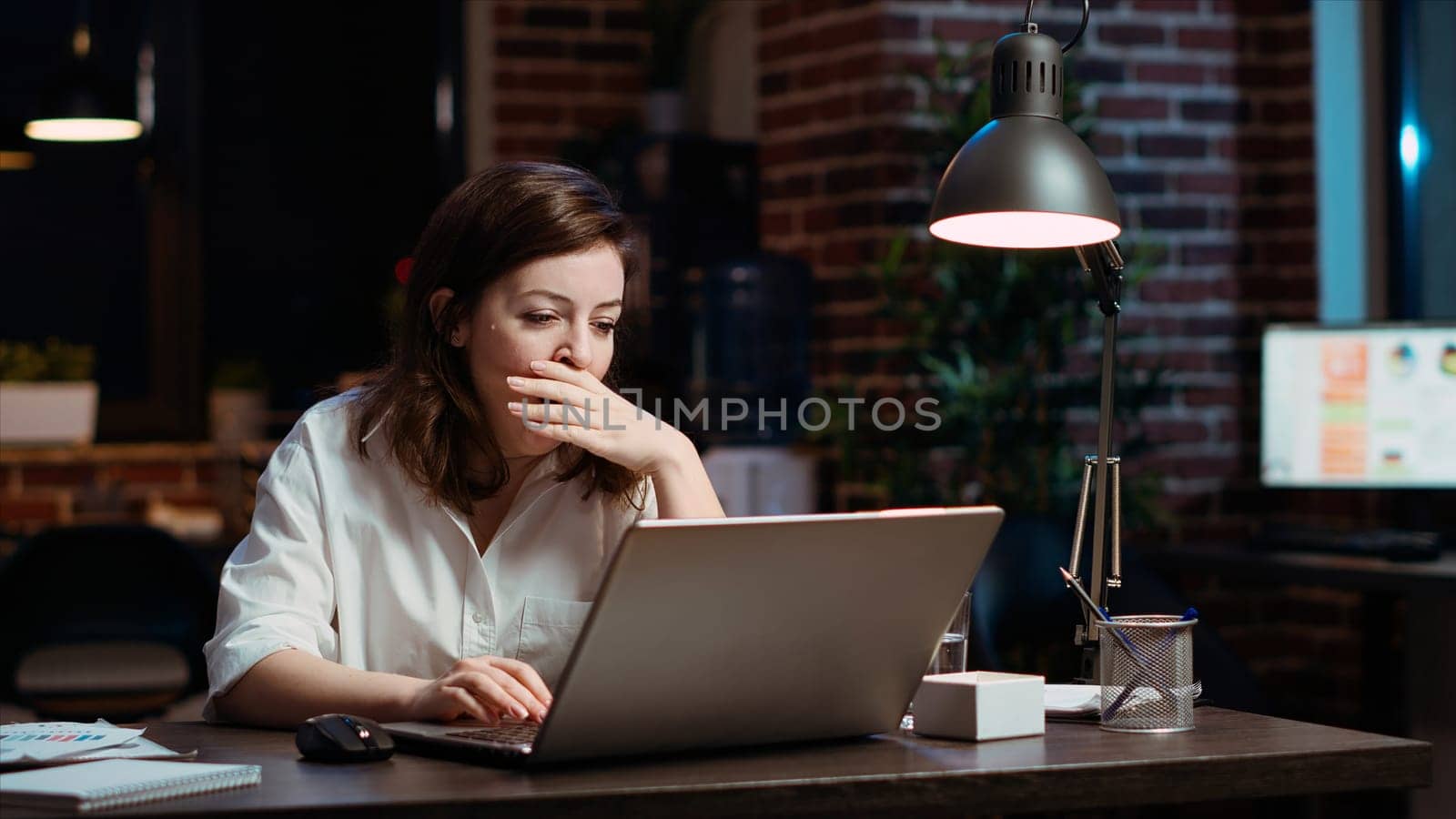 Weary accountant struggling to remain awake at workspace desk while inputting data on laptop, panning shot. Close up of exhausted businesswoman yawning in office while working overnight, camera B