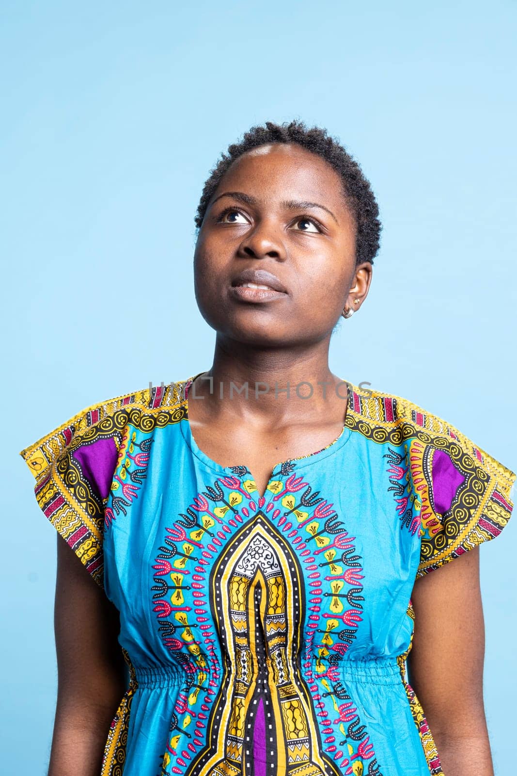 Portrait of serious person wearing traditional ethnic costume, woman posing on camera with confidence. African american girl looking gorgeous over blue background, feminine natural adult.