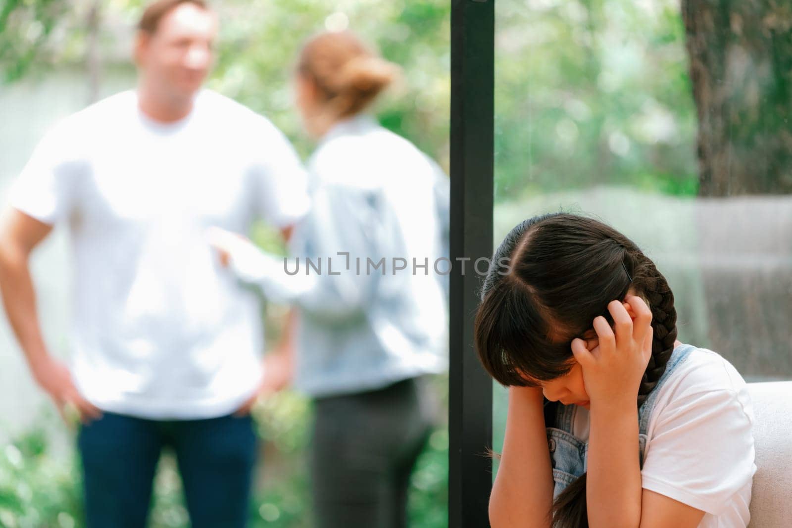 Stressed and unhappy young girl huddle in corner, cover her ears blocking sound of her parent arguing in background. Domestic violence at home and traumatic childhood develop to depression. Synchronos