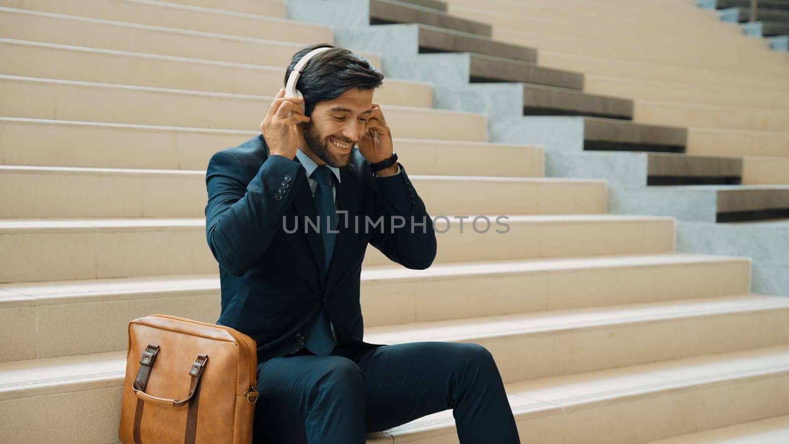 Professional business man picking and listening music by using headphone while sitting at stairs. Project manager dance while listening song while wearing headphone, suit. White background. Exultant.