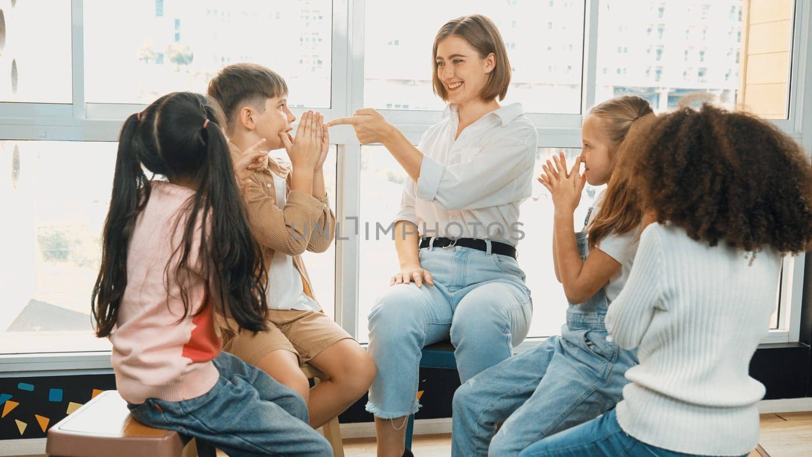 Smart caucasian teacher teaching while sitting on floor together surrounded by diverse students. Professional instructor playing with multicultural learner. Creative education concept. Erudition.